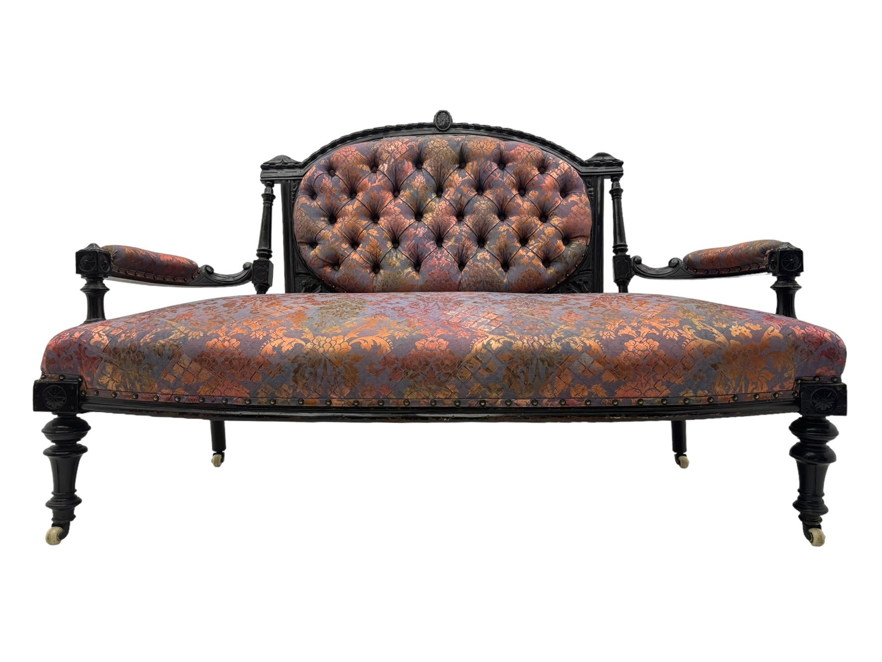 Late Victorian ebonised oak sofa, upholstered in muted purple fabric with iridescent floral pattern and nailhead trim, button-tufted backrest with carved crest rail and turned finials, scrolled padded arms supported by turned uprights, on turned supports with ceramic castors