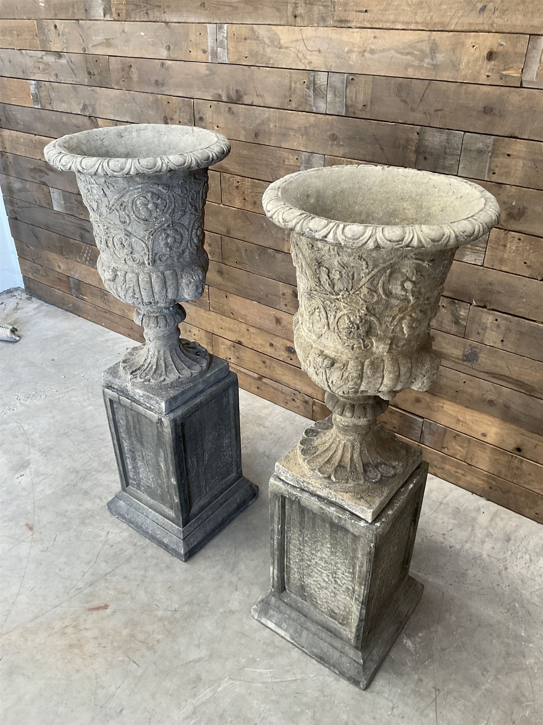 Pair of Georgian design cast stone garden urns, egg and dart rim with raised floral motifs, pedestal base terminating on square plinth, raised on square block plinth