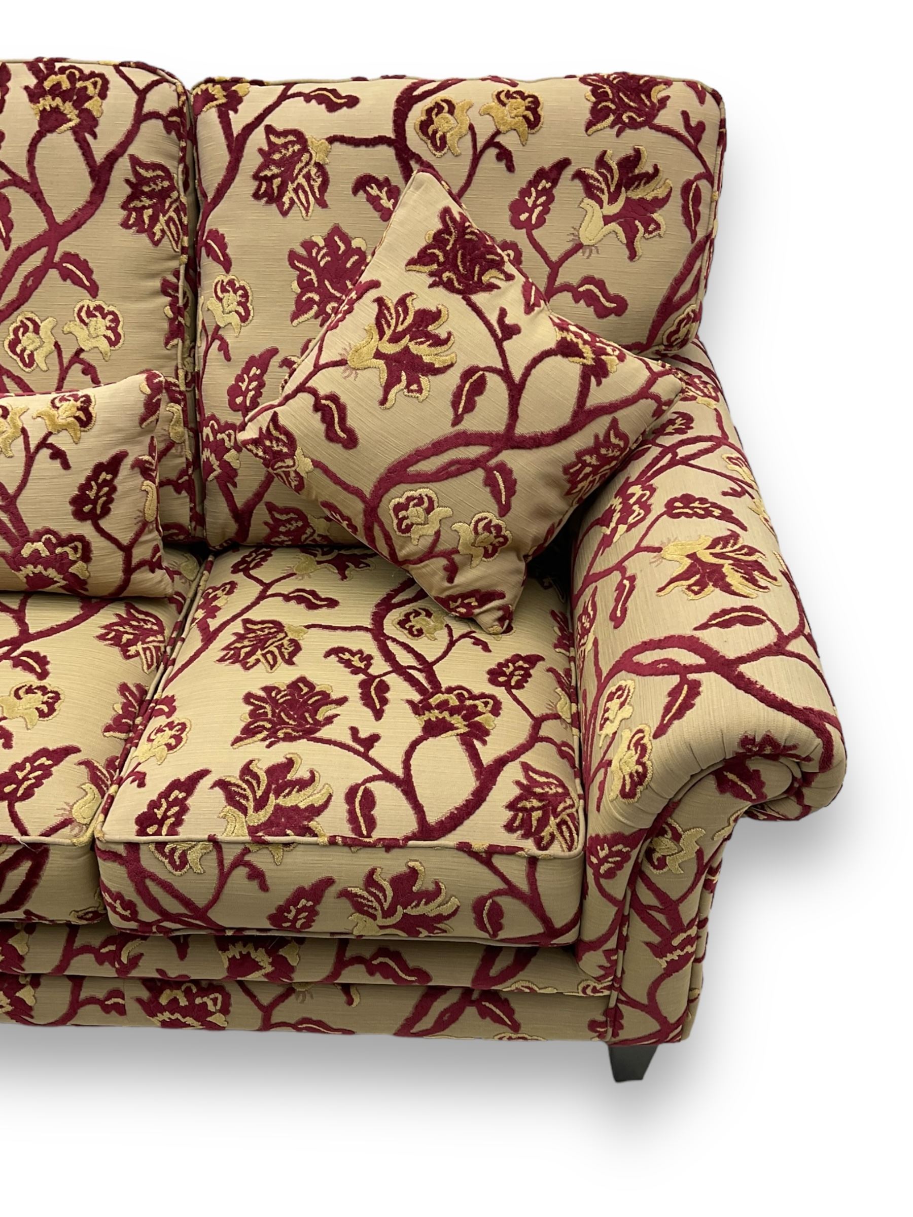 Contemporary upholstered three-seat sofa, scroll arms and loose cushions, embossed beige ground fabric with red and gold floral motifs, on tapered wooden supports