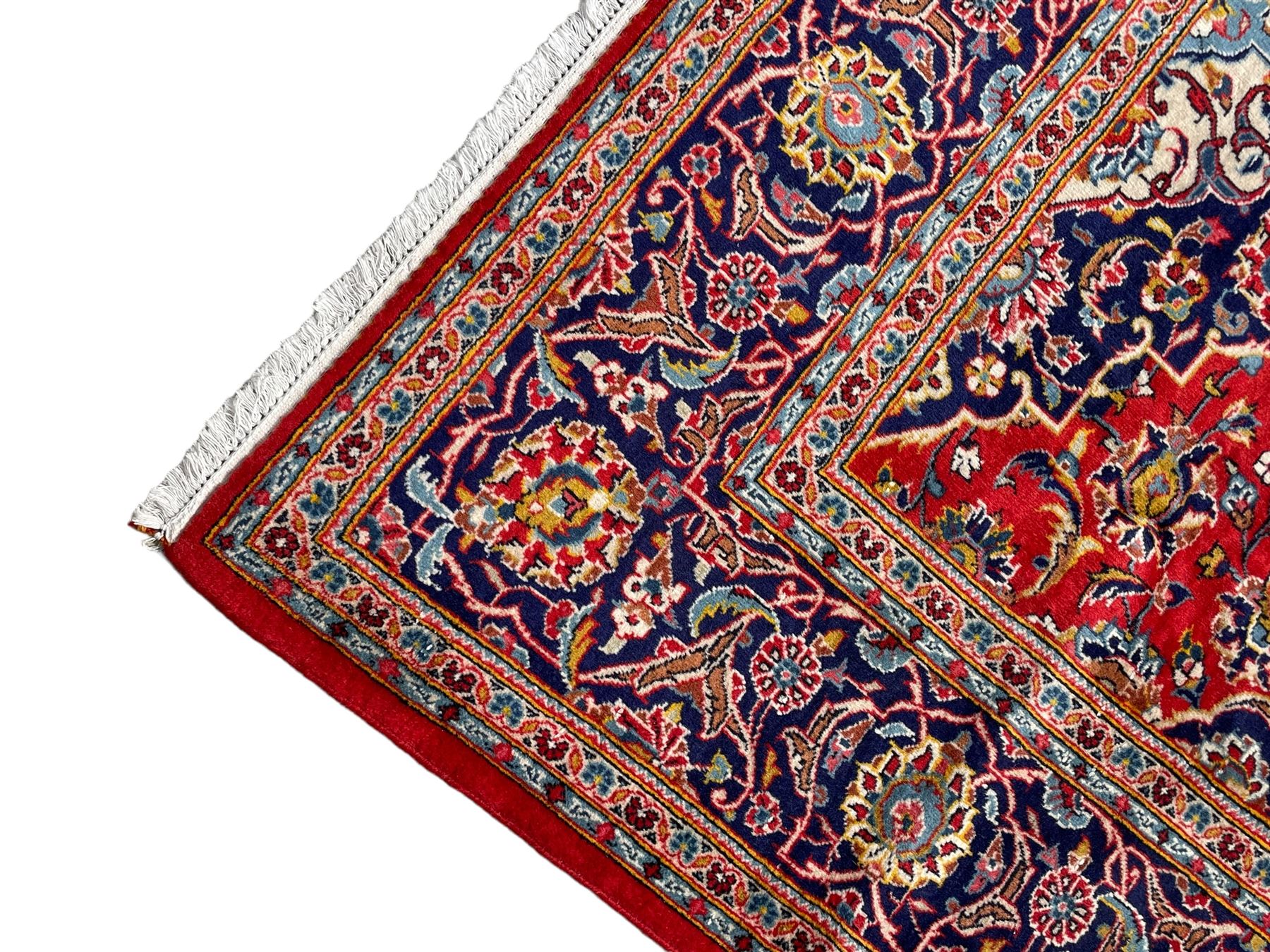 Persian Kashan crimson ground rug, overall arabesque design, the field decorated with swirled leafy branches and palmettes, indigo ground border decorated with repeating pattern, within guard stripes 