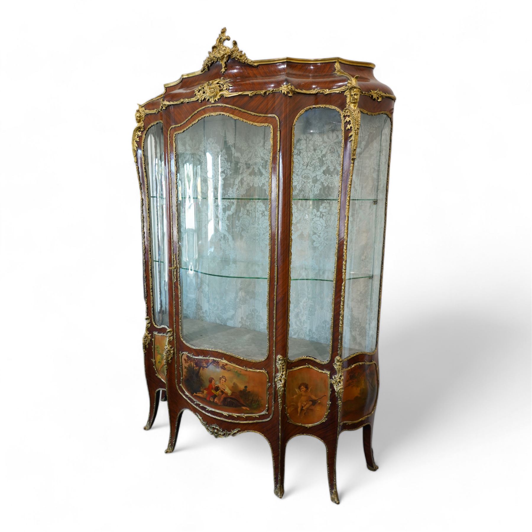 Large early 20th century French gilt metal mounted vitrine, serpentine and bombe form, C-scroll pediment decorated with extending flower heads, shaped front enclosed by single glazed door and glazed with shaped glass panes with foliate cast edging, floral pattern silk lined interior fitted with two glass shelves and one lined shelf, hand painted with Vernis Martin design panels depicting garden courting scene and putti, on cabriole supports mounted by shell and C-scroll mounts 