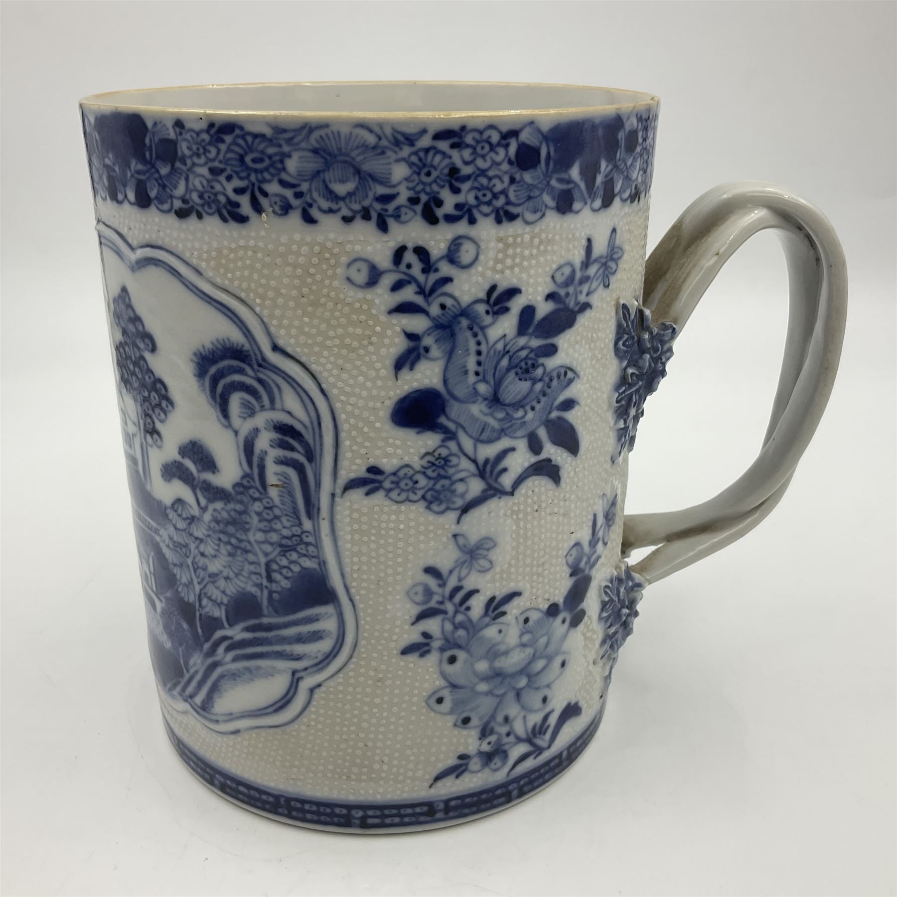 Two large 18th century Chinese blue and white tankards, each of cylindrical form, the first example with foliate mounted strap handle, the body painted with a shaped panel depicting a riverside landscape with pagodas and pine trees, against a textured ground decorated with floral sprays, H15cm D12cm, the second with serpent handle, the body painted with a similar riverside landscape, H14cm D12cm