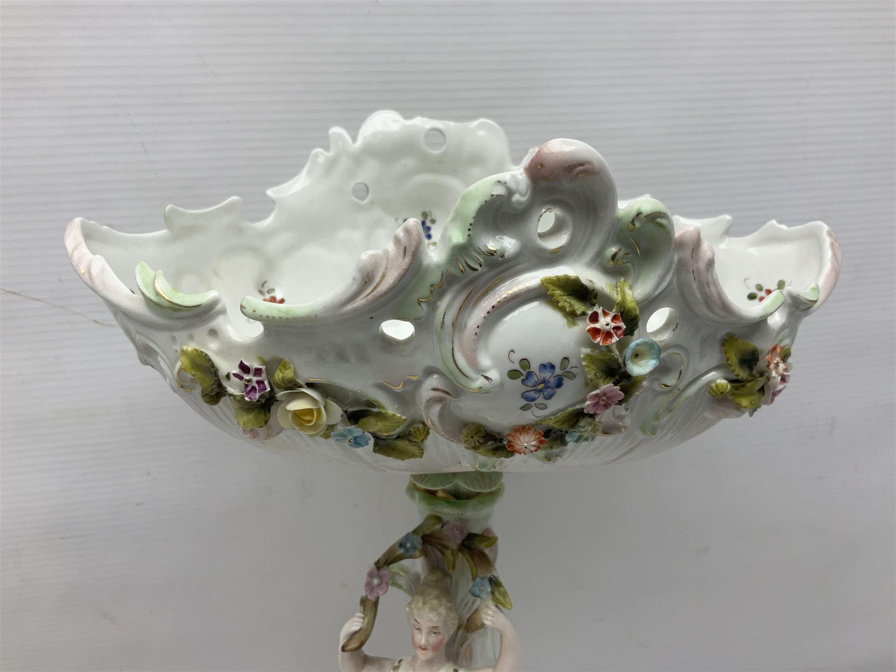 Early 20th century porcelain garniture, the central shaped basket upon a column supported by a female figure, and base with scroll feet, flanked by a pair of four branch candelabra, the scrolling branches and central stem leading to urn shaped sockets over foliate drip pans, upon shaped columns and bases with applied putto, the whole with encrusted flowers throughout, basket H33.5cm, candelabra H52cm