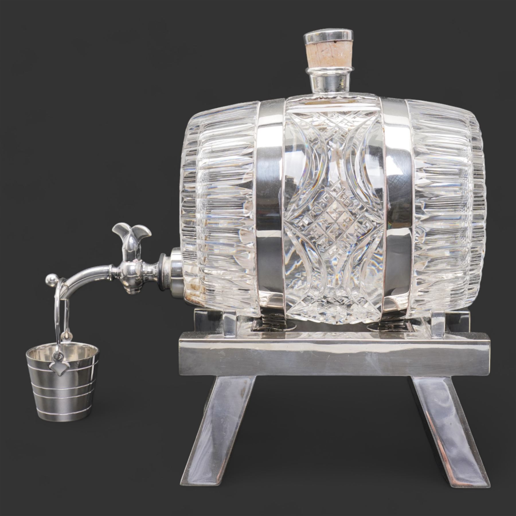 Late 19th/ early 20th century cut glass and silver plate whisky decanter by John Grinsell & Sons, of barrel form, with cork stopper and silver plate strapwork mounts, the tap suspending a bucket form measure, on trestle form support, H24cm x L24cm 