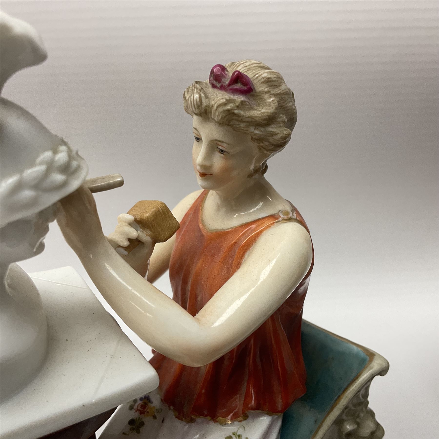 Pair of late 19th/early 20th century Naples figures modelled engaged in the arts, the first example modelled as a female figure seated before an easel, painting a figural scene, the second modelled as a female figure seated before a bust with scultping tools in hand, each upon an oval base with naturalistic painted ground, each with blue crowned N mark beneath, each approximately H16cm, including base L15xm
