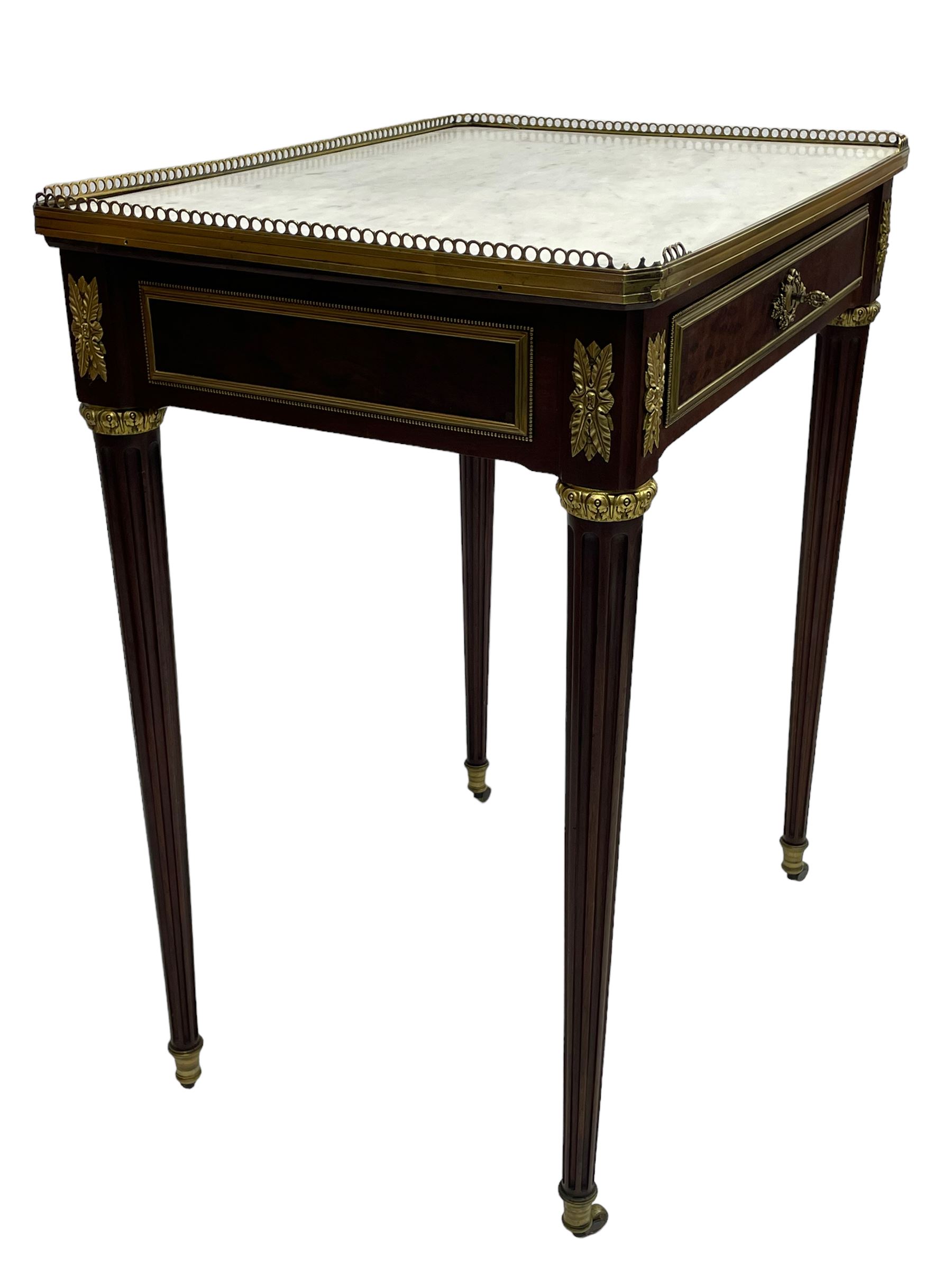 Late 19th century French figured plum pudding mahogany side table, canted rectangular white marble top with raised gilt metal gallery, fitted with a single frieze drawer, central extending floral cast ormolu escutcheon and applied moulded edging, on tapering turned and fluted supports with brass cups and castors, decorated with cast foliate mounts and foliate cast collars 