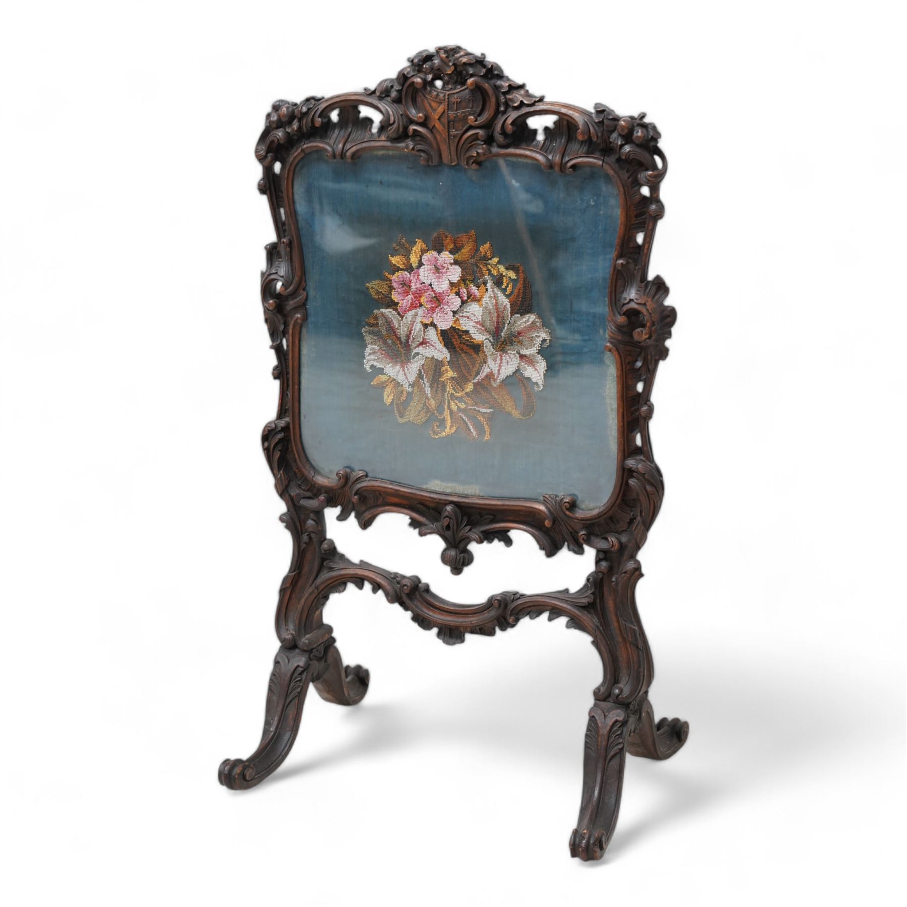 19th century carved wood fire screen, shield pediment decorated with oak leaves and acorns, the surrounding shell carved frame decorated with scrolling foliage, bead needle work panel depicting floral bouquet, on foliate carved splayed supports with scrolled terminals 