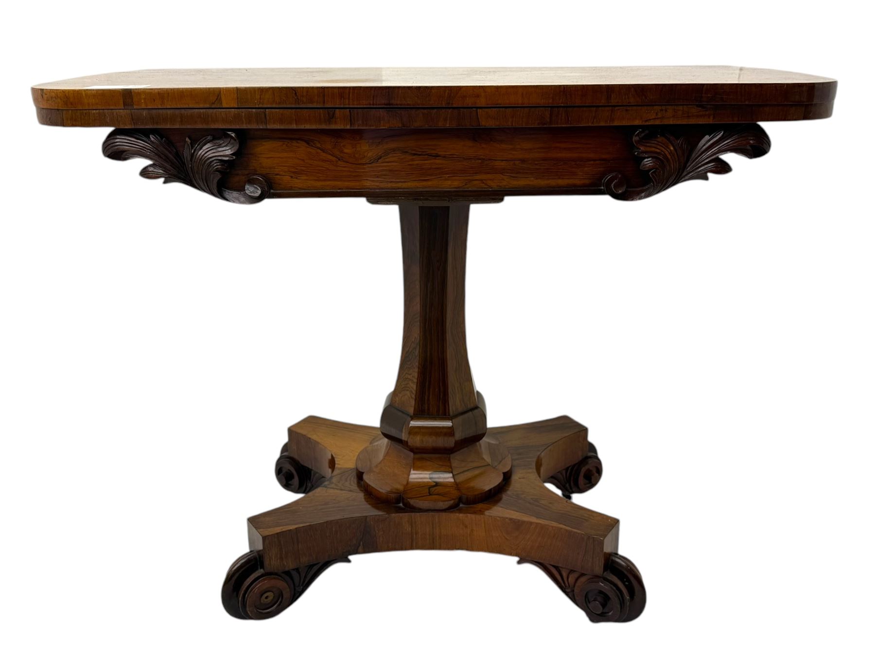 Early Victorian rosewood card table, rectangular foldover swivel top with rounded corners, red baize-lined surface, frieze adorned with carved foliate motifs, supported by a central octagonal waisted column on shaped quatrefoil base, terminating in scrolled feet with concealed metal castors