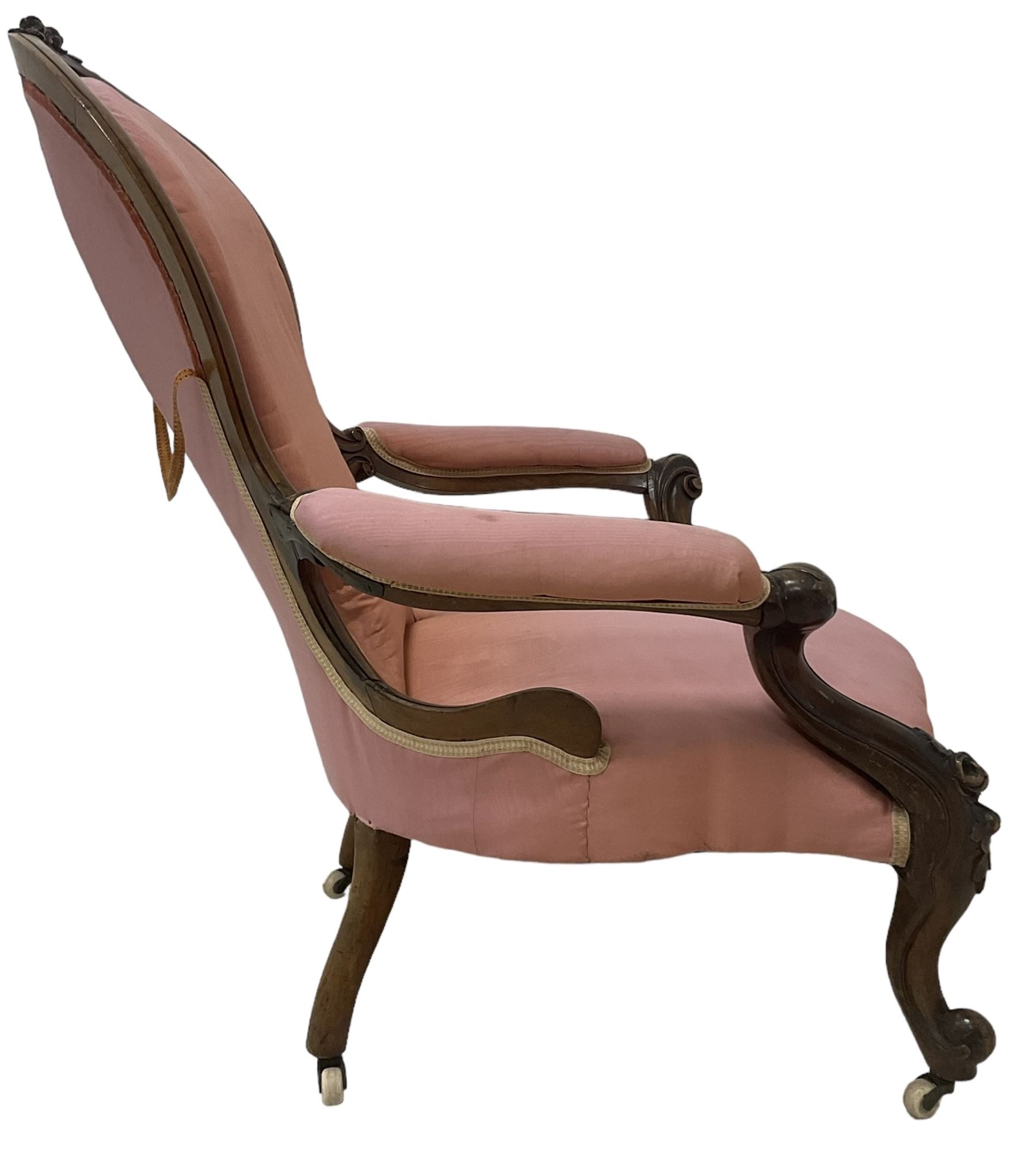 Victorian mahogany framed open armchair, spoon-back and sprung seat upholstered in pink fabric, scrolled arm terminals over serpentine fronted seat decorated with moulded carved floral motifs, cabriole supports with ceramic castors