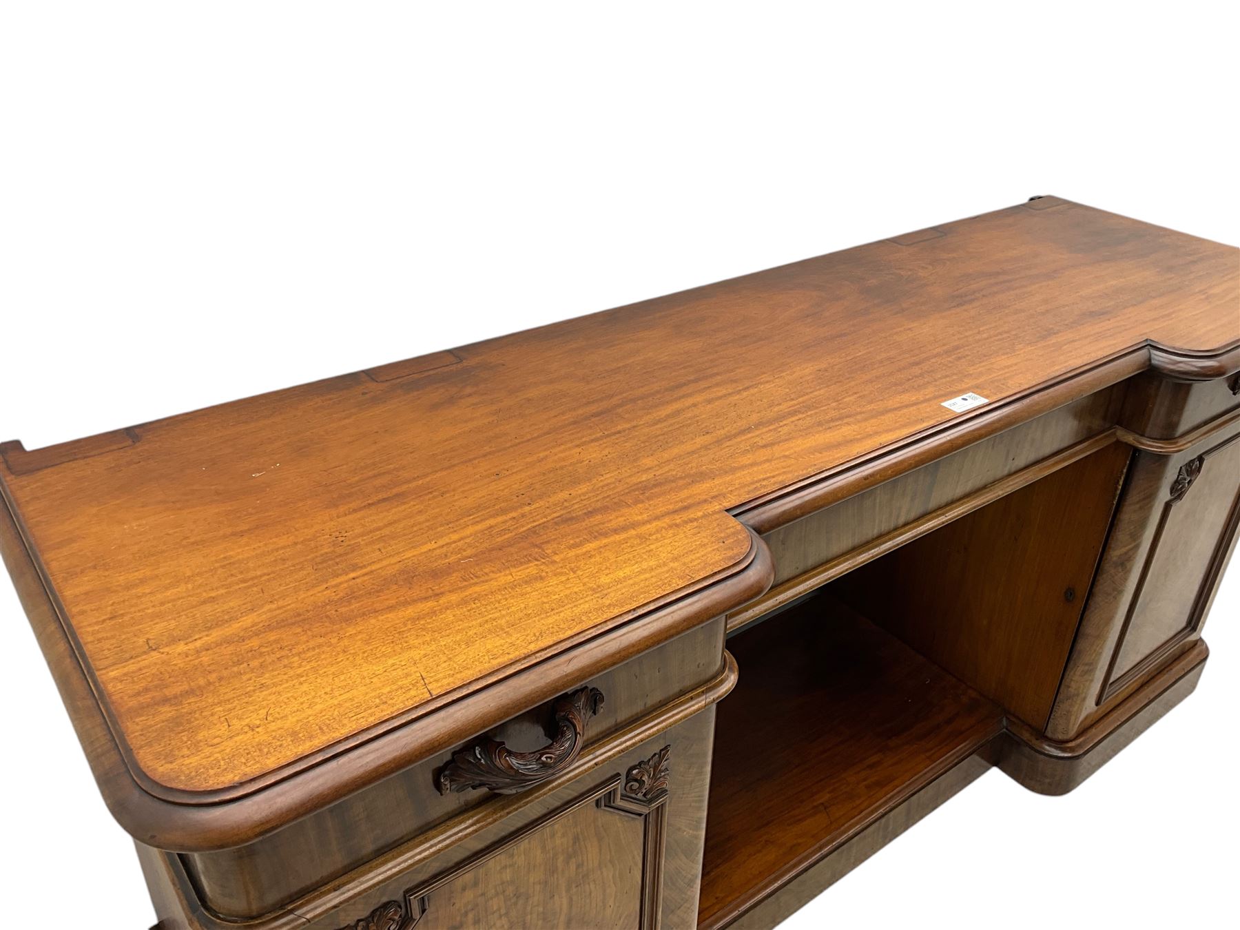 Mid-19th century figured mahogany sideboard, moulded reverse-break front, fitted with three drawers and two cupboards, carved faux bois handles, enclosed by panelled doors with book-matched veneers, applied geometric mouldings and scroll leaf decoration, on moulded plinth base 