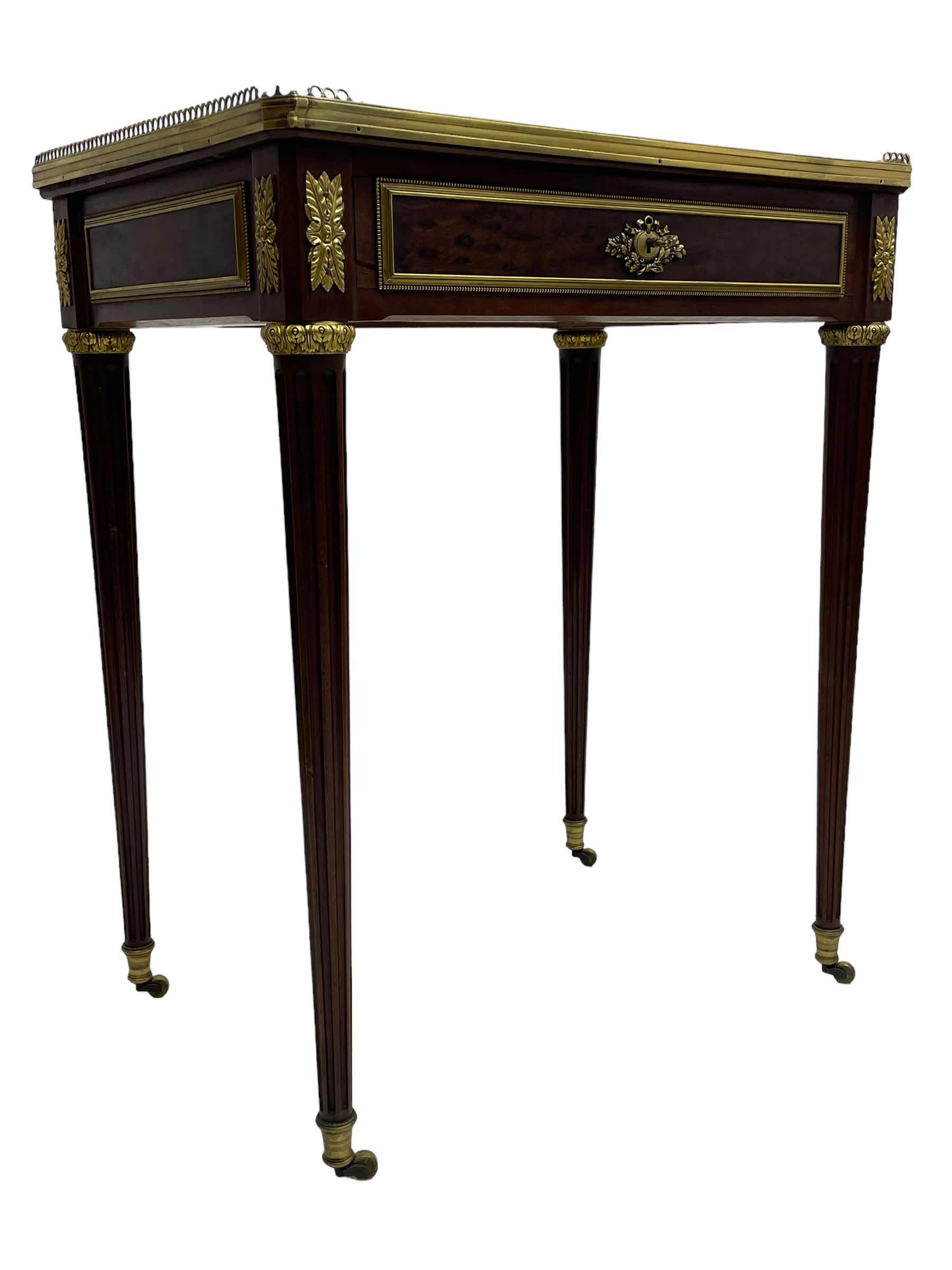 Late 19th century French figured plum pudding mahogany side table, canted rectangular white marble top with raised gilt metal gallery, fitted with a single frieze drawer, central extending floral cast ormolu escutcheon and applied moulded edging, on tapering turned and fluted supports with brass cups and castors, decorated with cast foliate mounts and foliate cast collars 