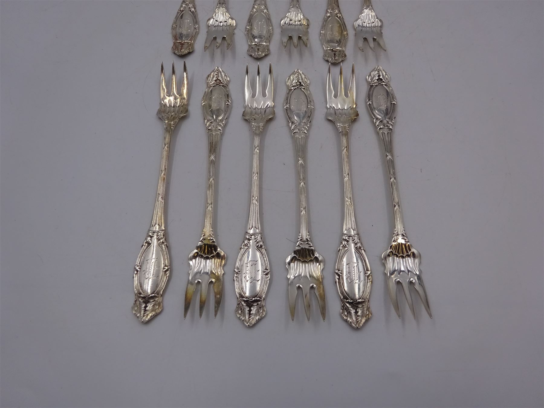 Set of twelve American silver oyster/pickle forks, three pronged forks, the stem decorated with ribbon banding and foliate border surrounding an engraved initial, stamped Sterling 