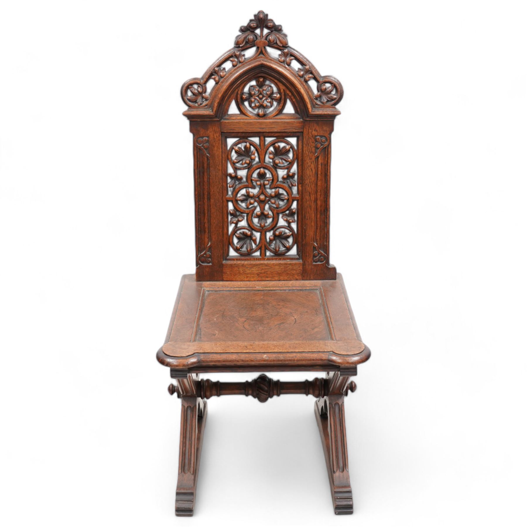 In the manner of Augustus Pugin - 19th century Gothic Revival oak hall chair, shaped pediment carved and pierced with foliate decoration, panelled back pierced with floral quatrefoil and scrolled decoration, panelled seat with moulded seat frame with projecting curved corners, on moulded X-framed supports united by turned and faceted stretcher, on sledge feet 