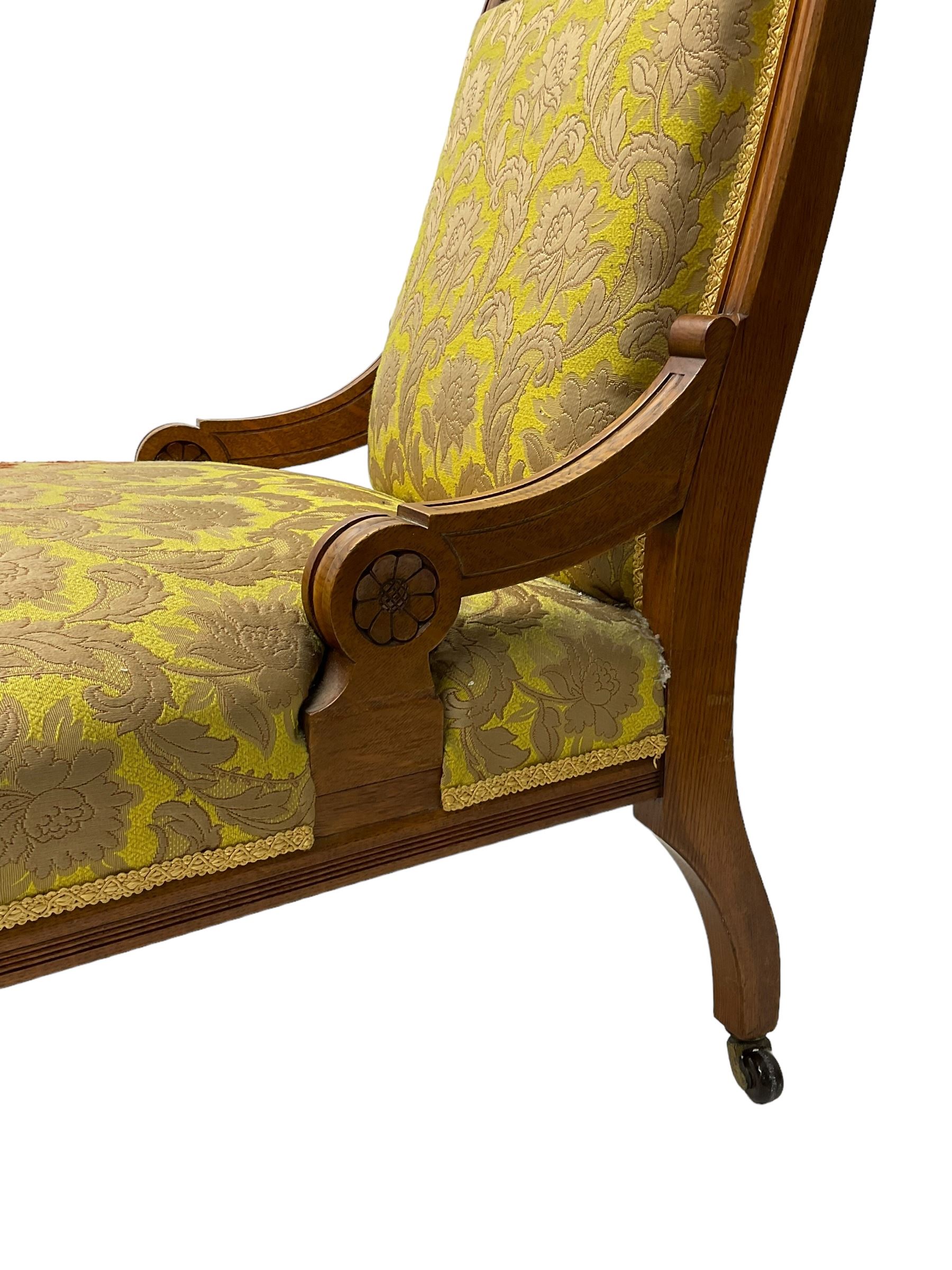 Late Victorian oak nursing chair, moulded frame with flower head carved roundels, upholstered in floral pattern fabric, on turned front feet with brass and ceramic castors 