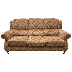 Parker Knoll - three-seat 'Oakham' hardwood-framed sofa, upholstered in red and gold floral pattern fabric, on turned feet with brass cups and castors 