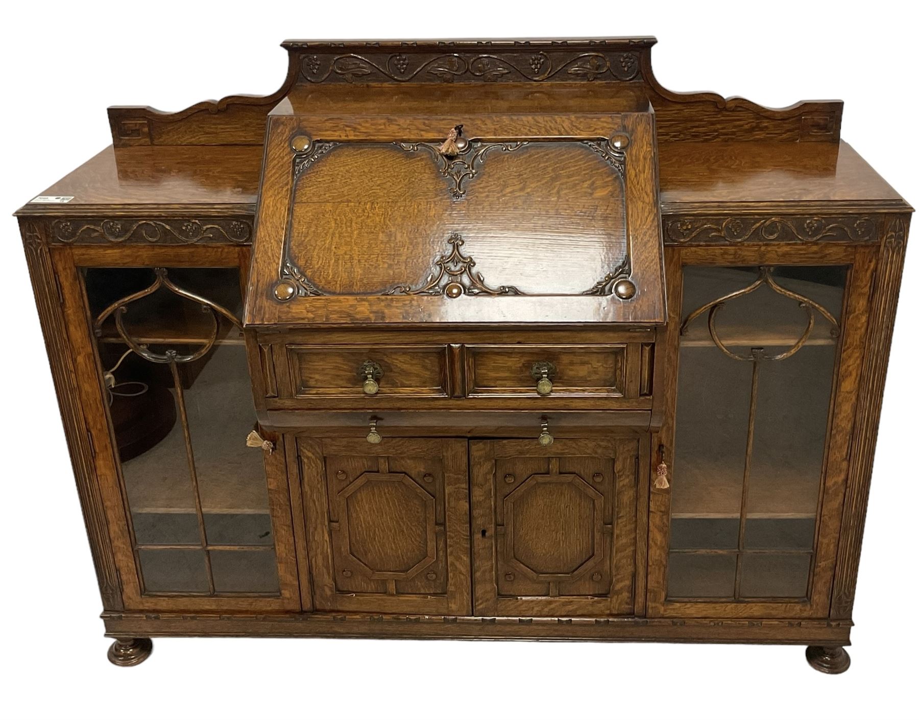 Early 20th century heavily carved oak bookcase bureau, raised back with grape-vine carved frieze, over fall-front enclosing fitted interior, above two short drawers and curved frieze drawer, flanked by astragal glazed and panelled cupboards, on turned feet