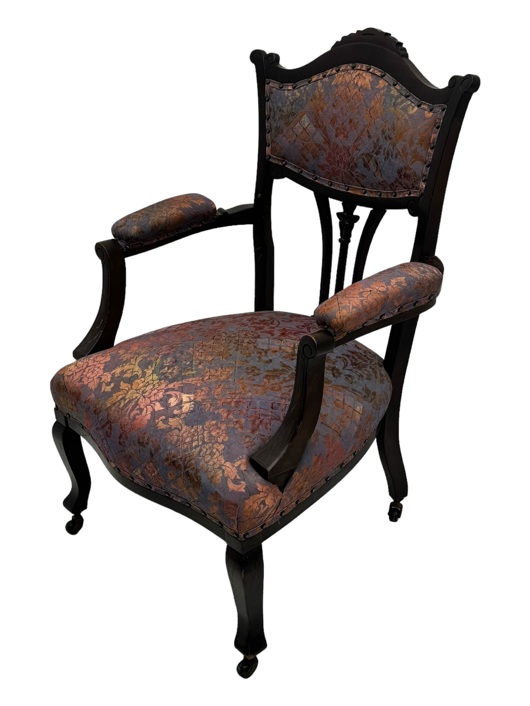 Late Victorian mahogany framed armchair, upholstered in muted purple fabric with iridescent floral pattern and nailhead trim, central foliate carved splat over padded scrolled arms, on cabriole supports fitted with castors