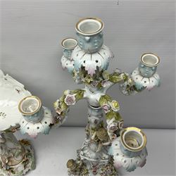 Early 20th century porcelain garniture, the central shaped basket upon a column supported by a female figure, and base with scroll feet, flanked by a pair of four branch candelabra, the scrolling branches and central stem leading to urn shaped sockets over foliate drip pans, upon shaped columns and bases with applied putto, the whole with encrusted flowers throughout, basket H33.5cm, candelabra H52cm