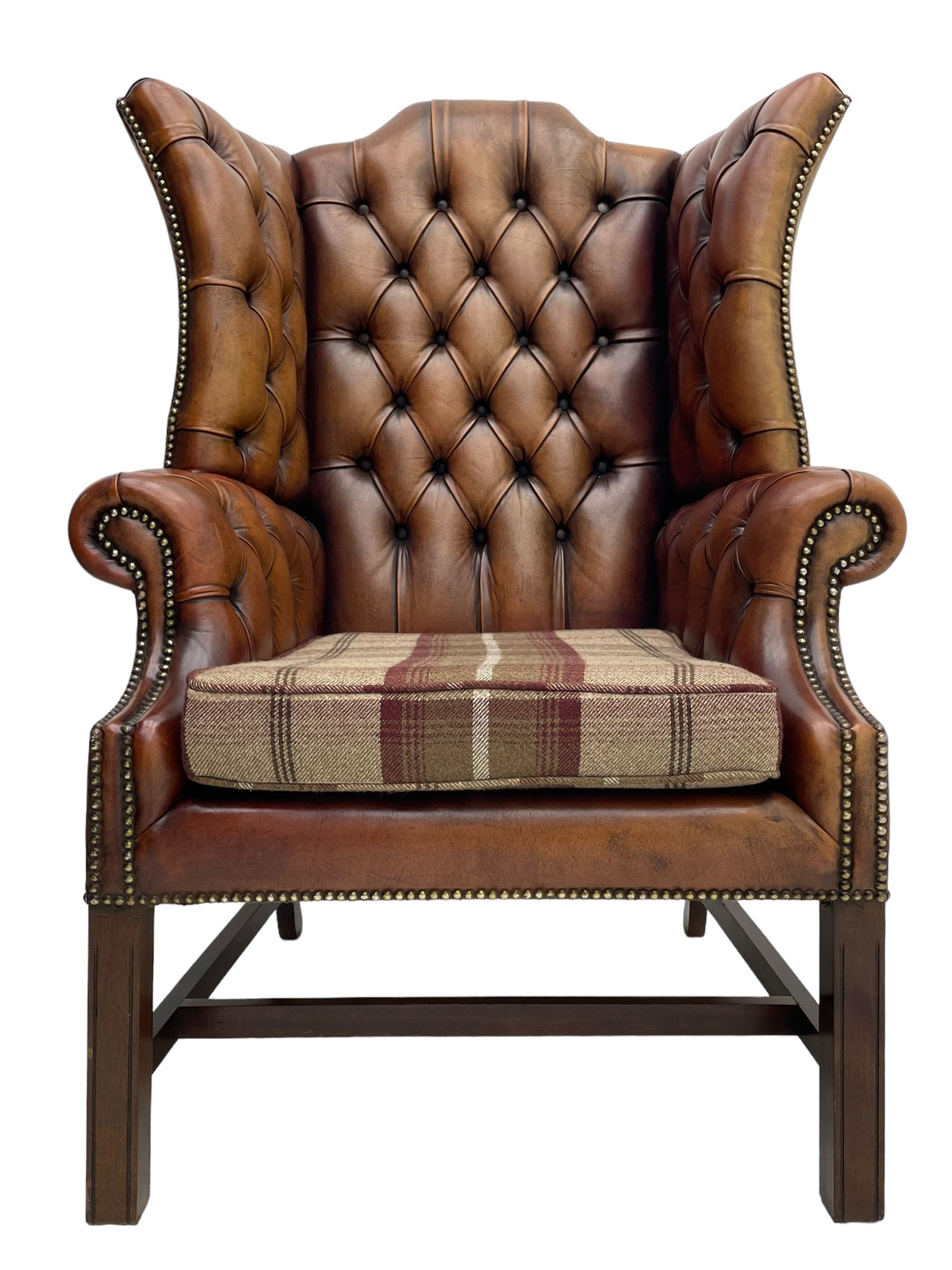 Georgian design hardwood-framed wingback armchair, shaped cresting rail and deep wingback over rolled arms, upholstered in brown buttoned leather with studwork bands, the seat cushion upholstered in checkered fabric, on square supports united by stretchers (W86cm, H118cm, D86cm); together with matching rectangular footstool (63cm x 43cm, H37cm) 