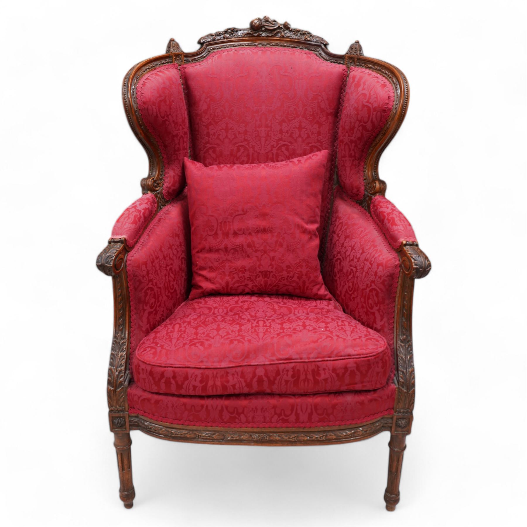 Late 19th century walnut framed fauteuil armchair, arched cresting rail carved with musical instruments and extending floral decoration, curved wing back with moulded and bead carved frame, upholstered in crimson fabric with repeating foliate pattern, acanthus carved arm terminals over acanthus and ribbon carved arm supports, the seat rail carved with fruiting foliage band, on turned and stop-fluted feet 