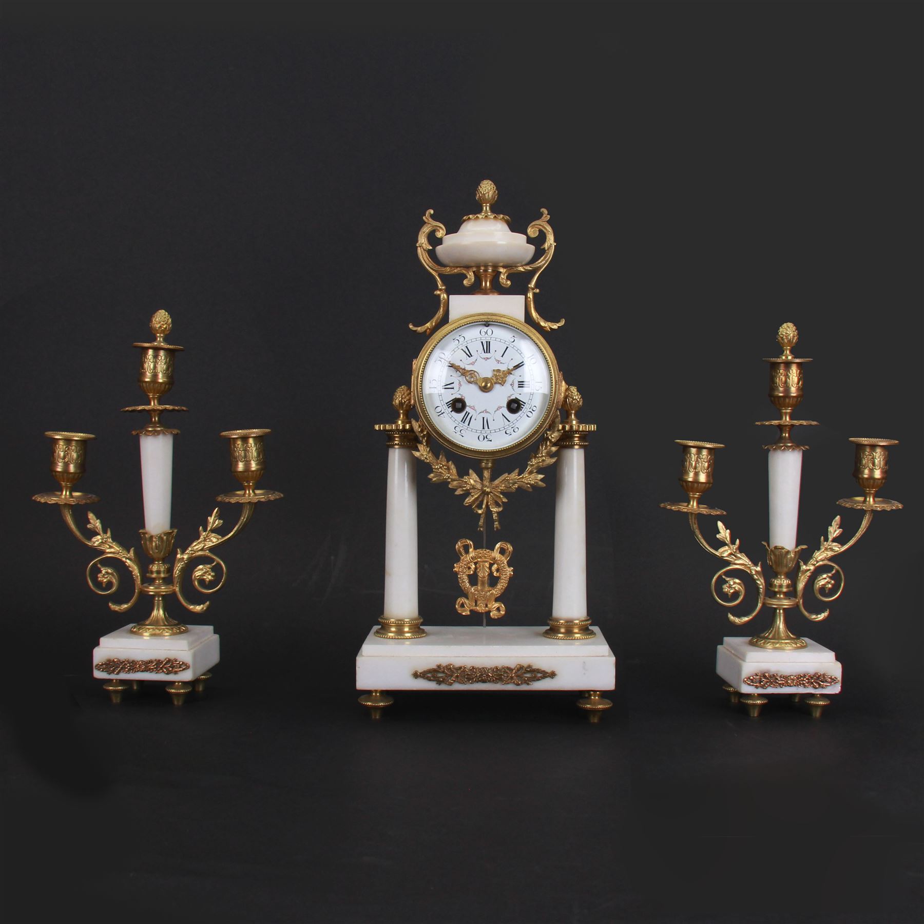 French - Louis XV style white marble and gilt mounted 8-day portico clock garniture c1905, drum movement surmounted by an oval shaped urn, supported on a pair of tapered column supports and raised on a rectangular plinth base, white enamel dial with floral garlands and Arabic numerals, Louis XV style gilt hands within a glazed bezel, twin train countwheel striking movement, striking the hours and half hours on a bell, with a pair of conforming three light candelabra, decorative matching pendulum and key.