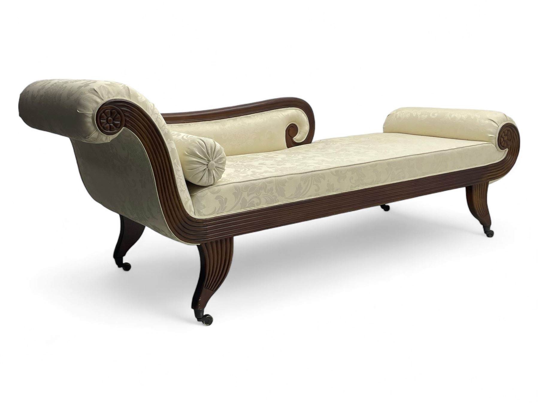 Regency design mahogany chaise longue, scrolled arms with carved rosette details, upholstered in cream damask fabric with bolster cushion, reeded frame supported by turned legs on brass castors