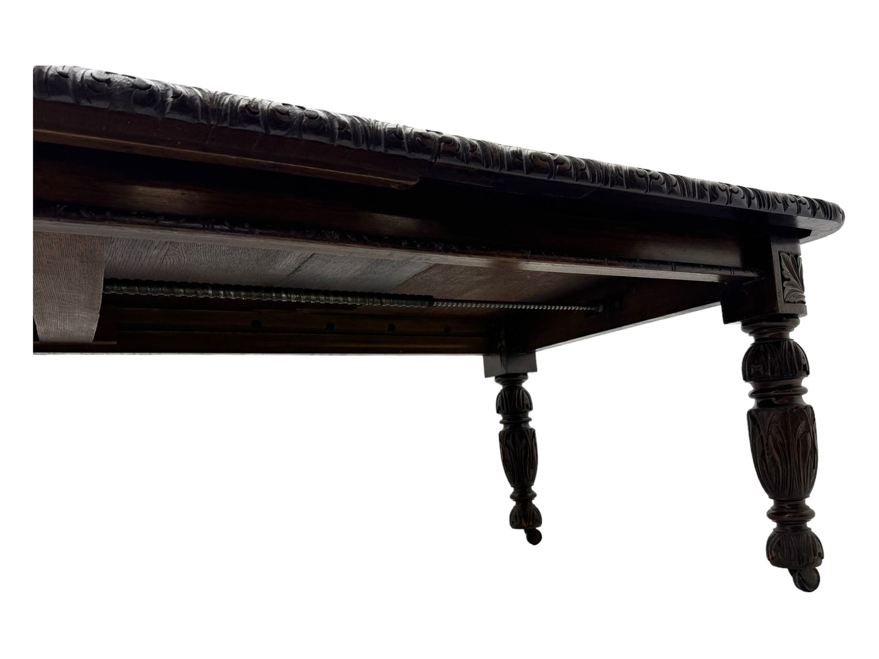 19th century heavily carved oak extending dining table, rectangular top with rounded corners and carved gadrooned edge, extending via winding mechanism with two additional leaves, raised on acanthus leaf-carved baluster supports terminating in ceramic castors