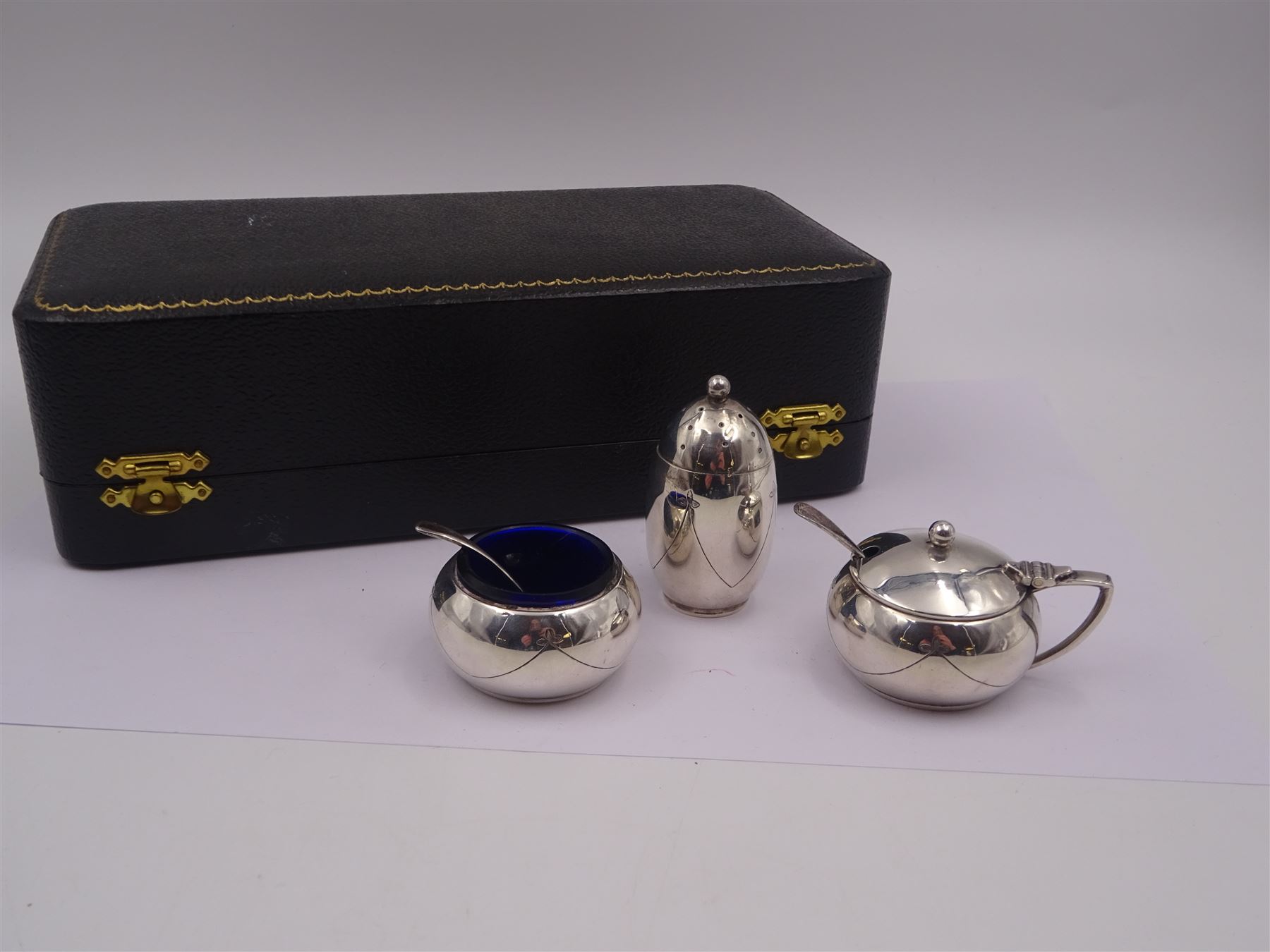 1950s silver three piece cruet set, comprising pepper shaker, open salt, mustard pot and cover, engraved with ribbon swags, hallmarked Joseph Gloster Ltd, Birmingham 1956, salt and mustard pots with blue glass liners, together with two condiment spoons, hallmarked, all within a fitted silk and velvet lined case 