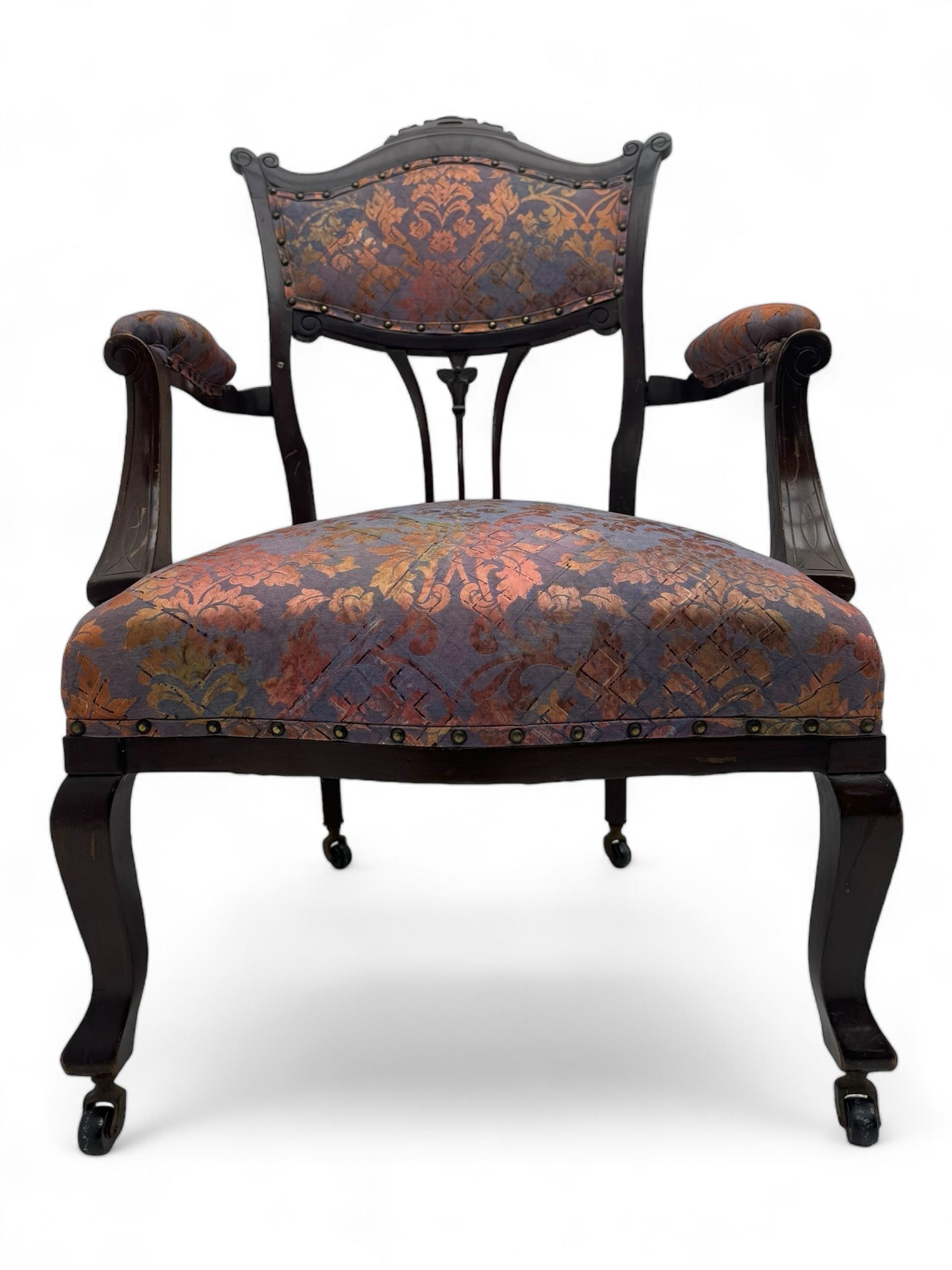 Late Victorian mahogany framed armchair, upholstered in muted purple fabric with iridescent floral pattern and nailhead trim, central foliate carved splat over padded scrolled arms, on cabriole supports fitted with castors