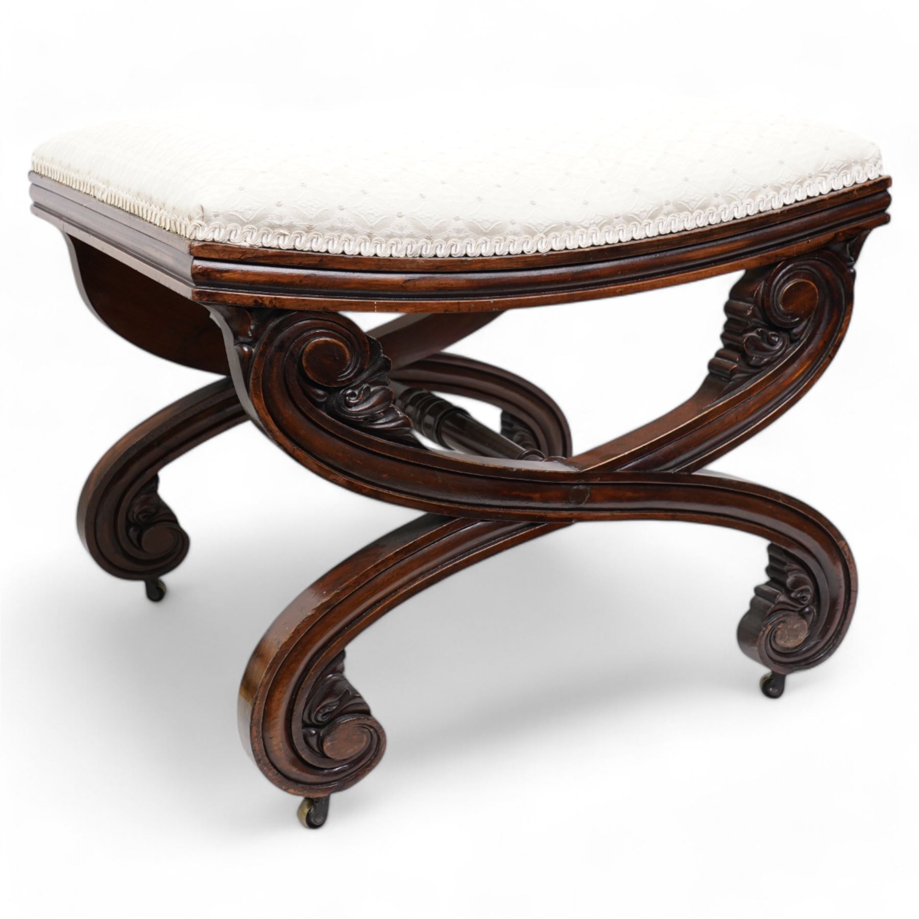 Early 19th century mahogany stool, the dished seat upholstered in cream fabric with repeating pattern, moulded seat rails, on curved X-frame supports carved with scrolls and foliage, united by turned stretcher, on brass castors