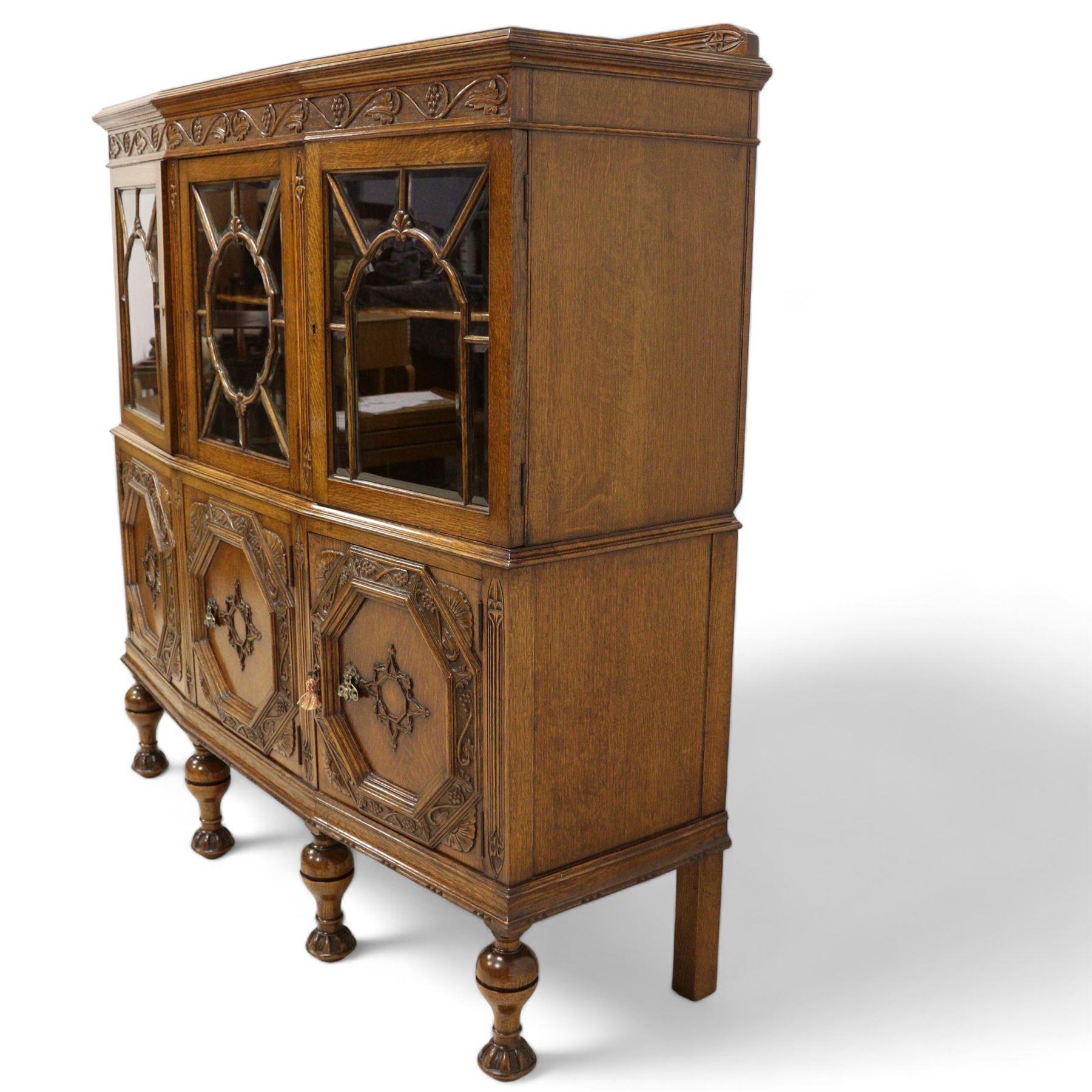 Early 20th century heavily carved oak display cabinet, frieze carved with grape vines over three astragal glazed doors with bevelled glass, over three cupboards applied with octagonal carved panels with further vine carvings, on turned and lobe carved supports