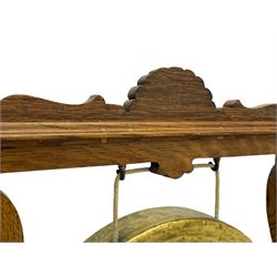 Early 20th century oak gong stand with gong and hammer, shaped cresting rail over moulded upper edge on shaped supports, hanging brass drum gong over fretwork middle rail, on raised platforms terminating to splayed feet