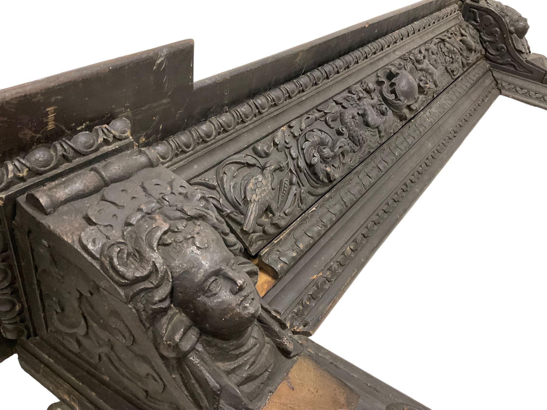 19th century Elizabethan design carved oak fire surround, reverse break-front lintel over egg and dart moulded upper edge, the ovolo-moulded frieze carved with scrolling leafy branches, central cartouche and putti riding dolphins, the uprights carved with female masks over carved busts, square tapering pilasters carved with ribbon tie over trailing fruit, on stepped moulded base 