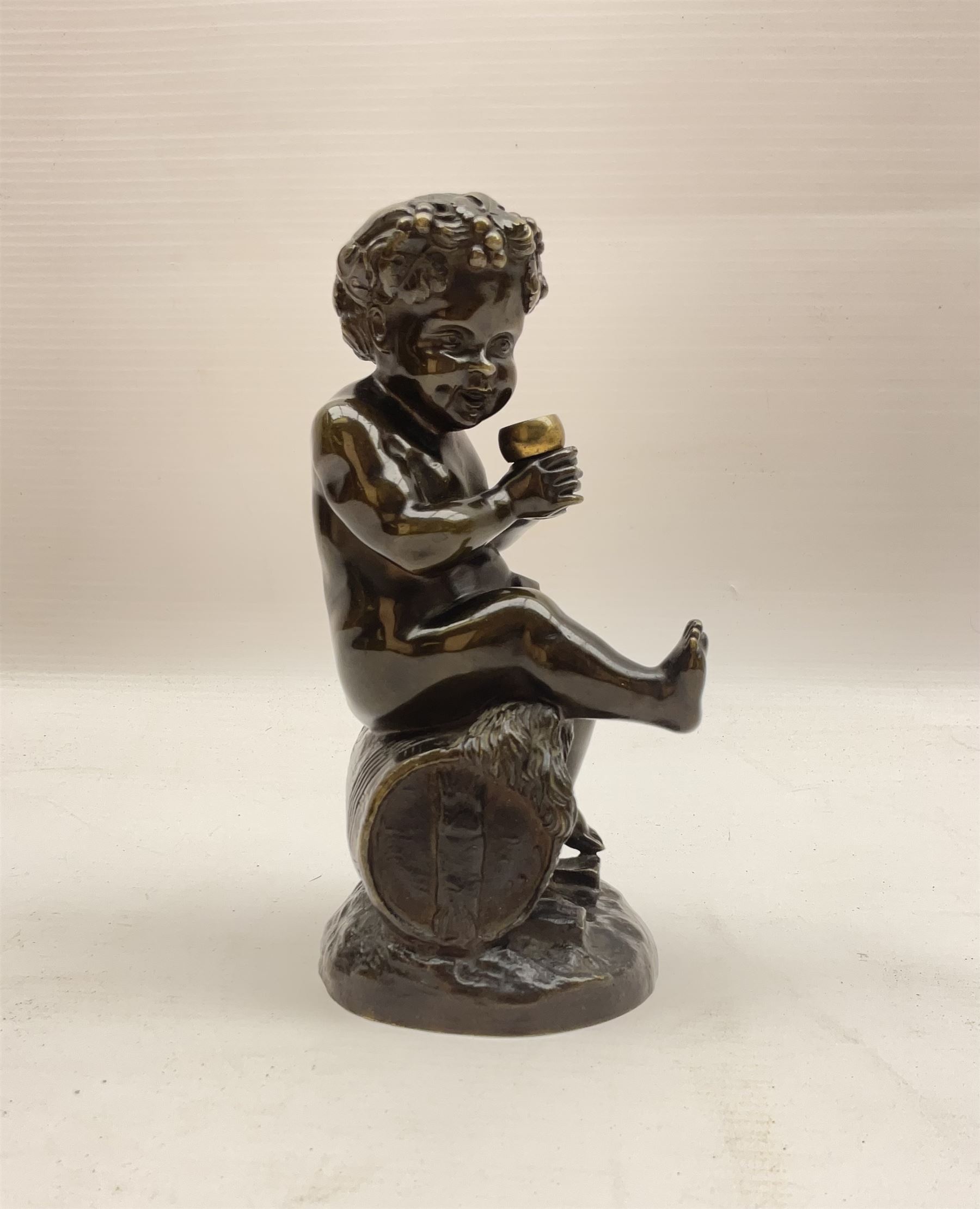 Bronzed metal model of a putti seated upon a barrel with goblet in hands, and fruiting vines adorning their head, H17.5cm