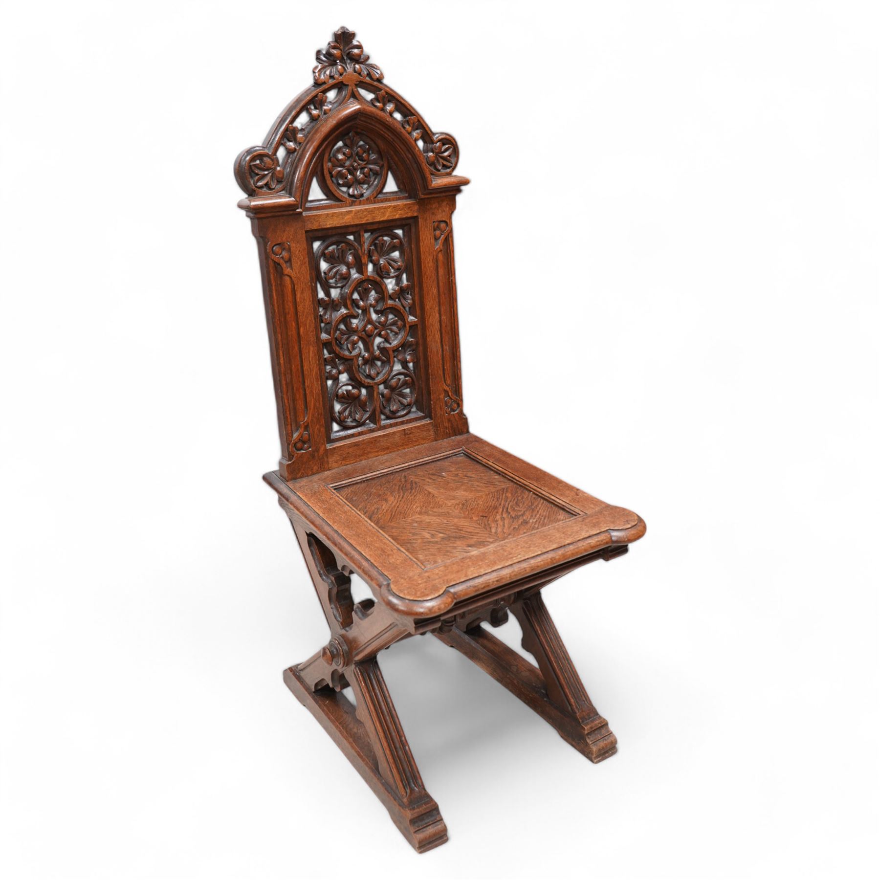 In the manner of Augustus Pugin - 19th century Gothic Revival carved oak hall chair, the pierced and carved arched pediment pierced and carved with scrolling foliage, the pierced back panel carved with a central pierced quatrefoil and leafage, panelled and eared seat over an architectural X-frame base with chamfered trefoil carvings and sledge feet