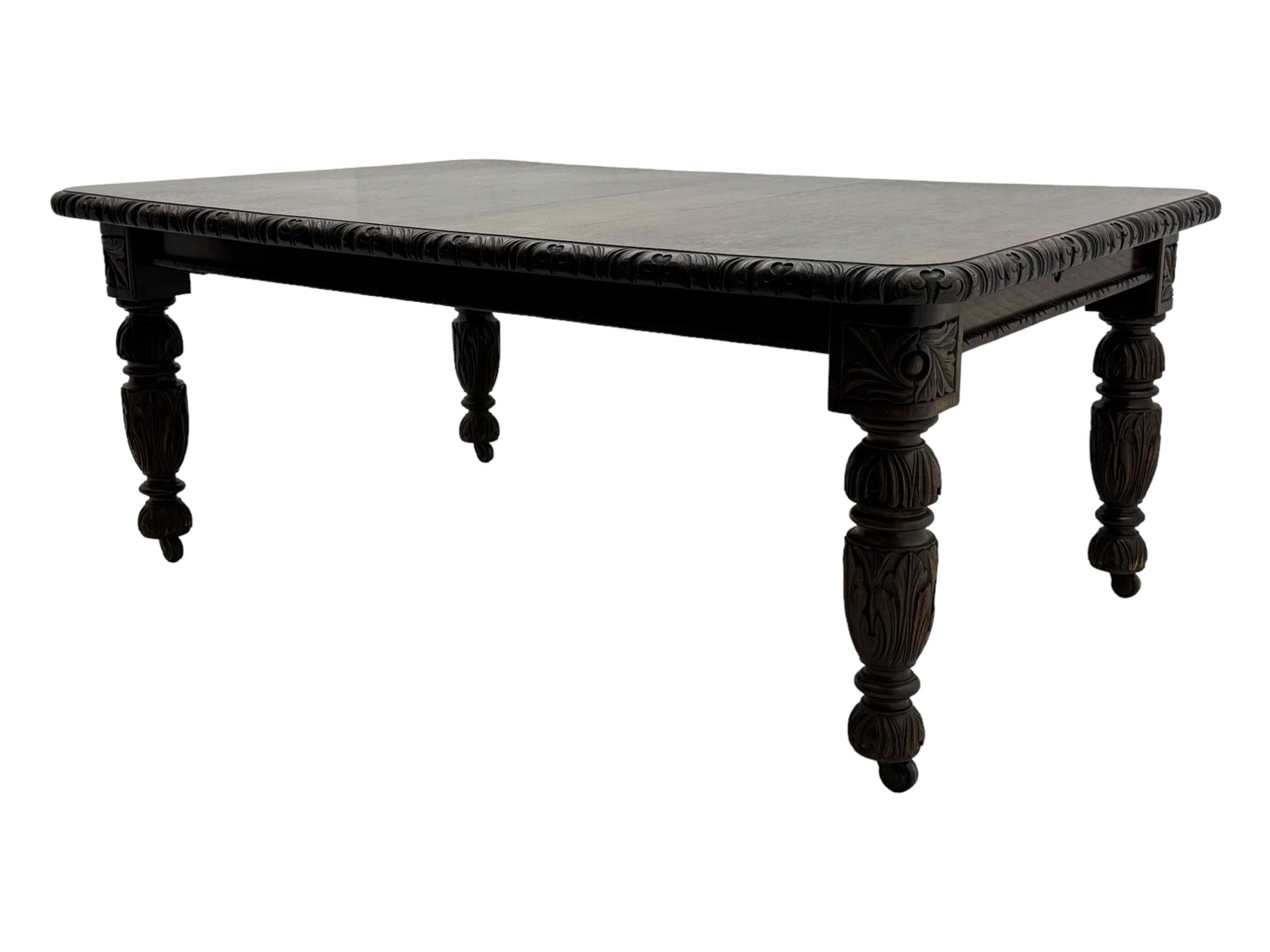 19th century heavily carved oak extending dining table, rectangular top with rounded corners and carved gadrooned edge, extending via winding mechanism with two additional leaves, raised on acanthus leaf-carved baluster supports terminating in ceramic castors