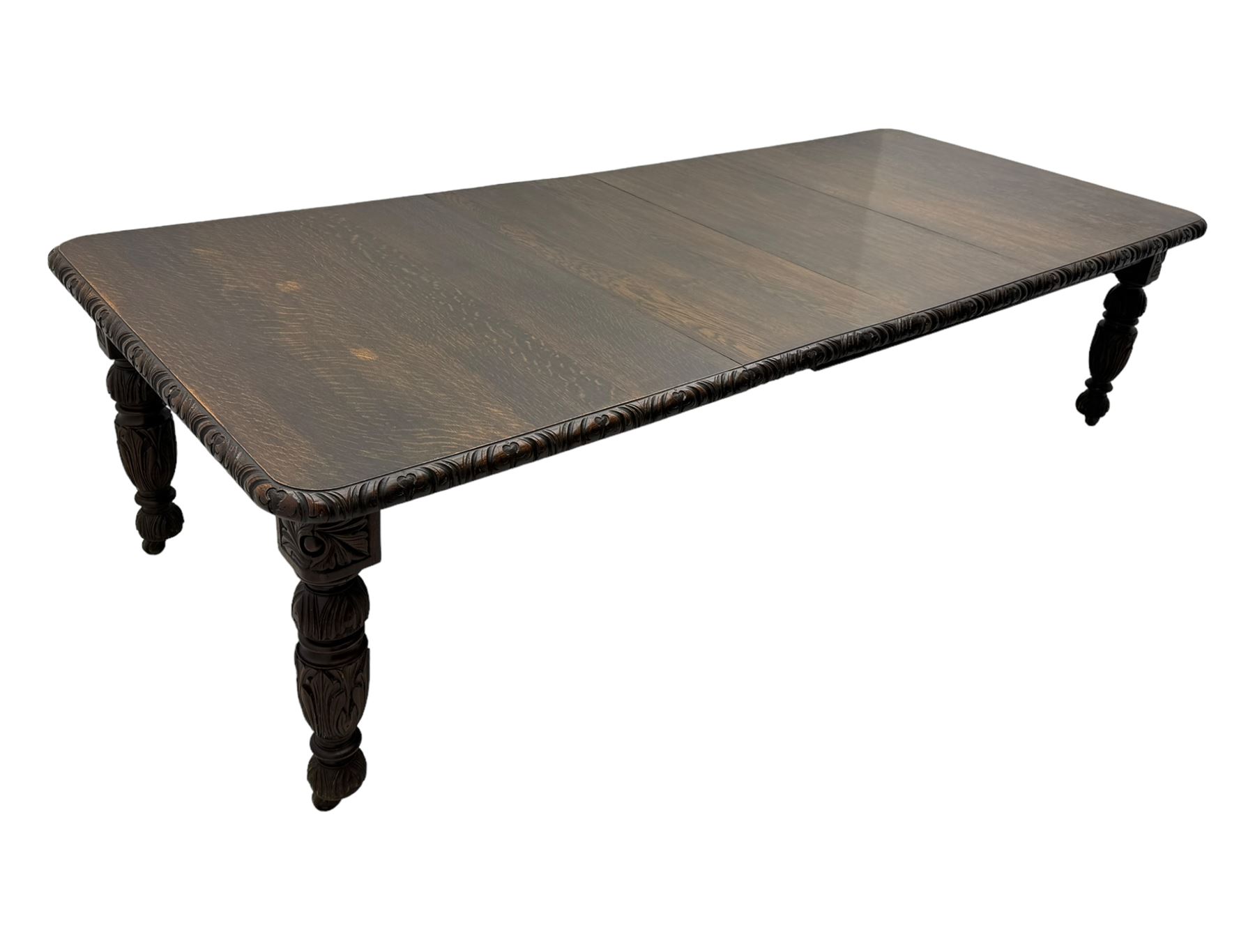 19th century heavily carved oak extending dining table, rectangular top with rounded corners and carved gadrooned edge, extending via winding mechanism with two additional leaves, raised on acanthus leaf-carved baluster supports terminating in ceramic castors