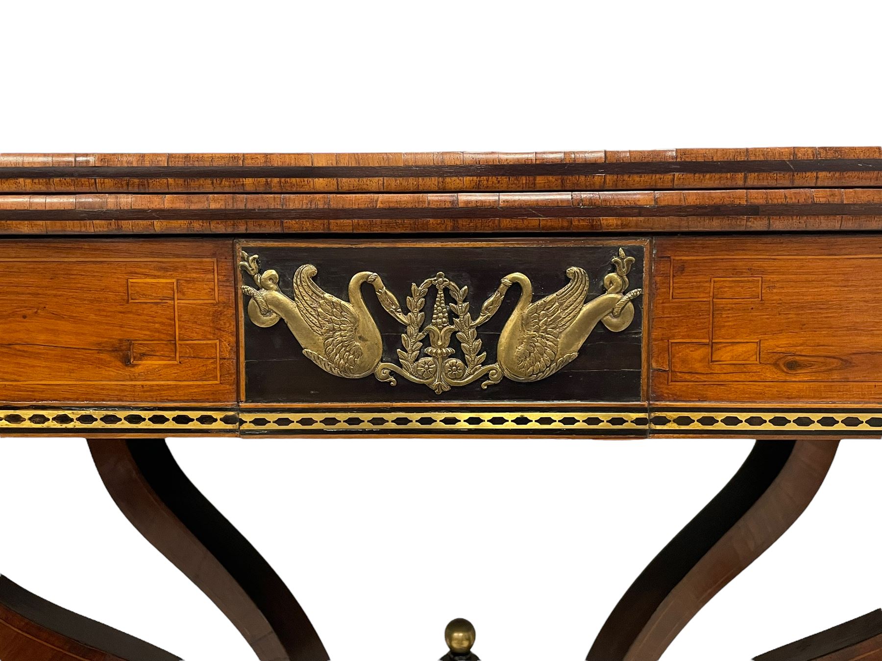 Regency inlaid rosewood card table, rectangular swivel and fold-over top with rounded corners and baize lined interior, the frieze decorated with a central ebony panel with applied gilt metal swan motifs, over a brass inlay band, raised on S-scroll supports united by concave platform base, terminating to out-splayed supports with brass cups and castors