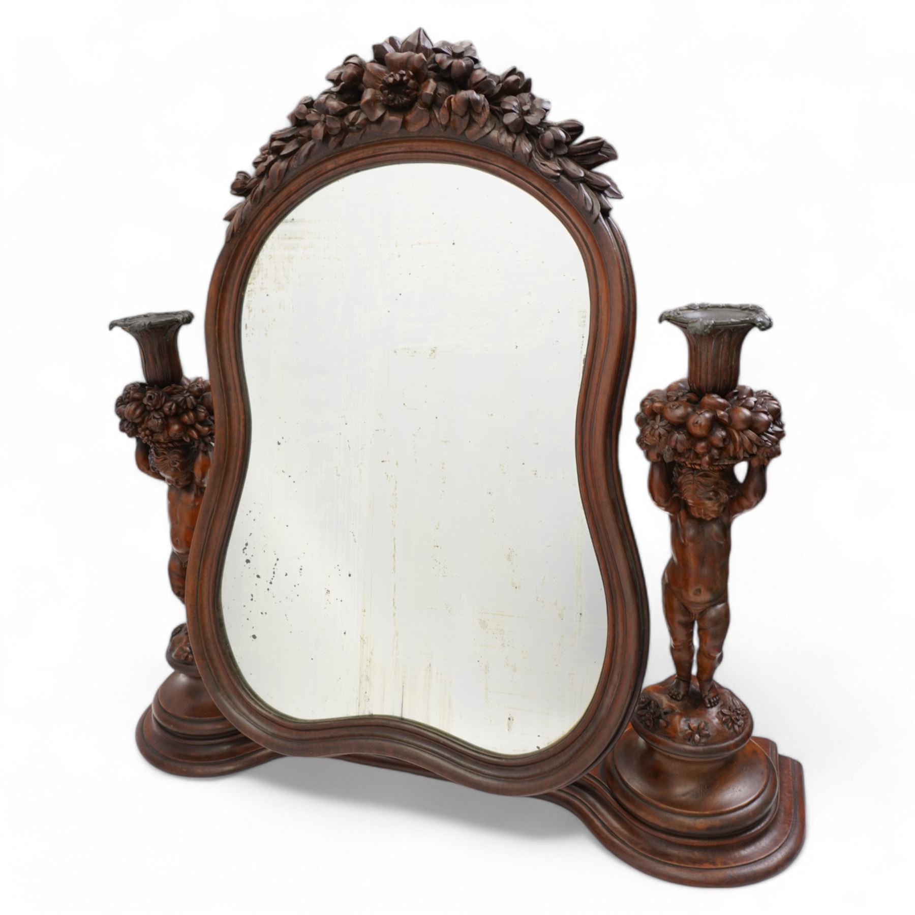 Late 19th century Northern European carved walnut dressing table mirror, the moulded arch top frame carved with flower heads and foliage, flanked by two putto holding baskets of flowers and fruit with candle sconces, on shaped moulded base, plain mirror plate 