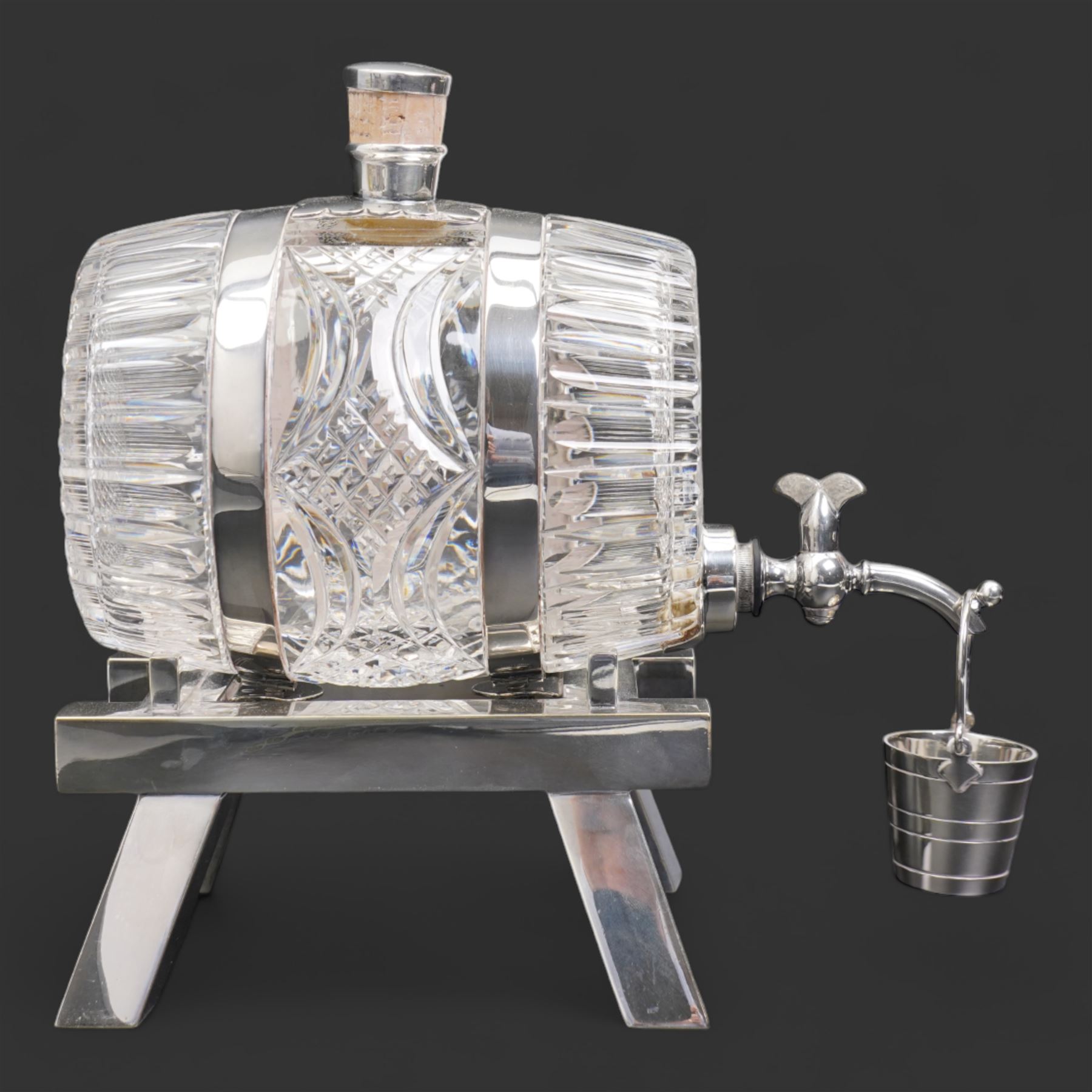 Late 19th/ early 20th century cut glass and silver plate whisky decanter by John Grinsell & Sons, of barrel form, with cork stopper and silver plate strapwork mounts, the tap suspending a bucket form measure, on trestle form support, H24cm x L24cm 