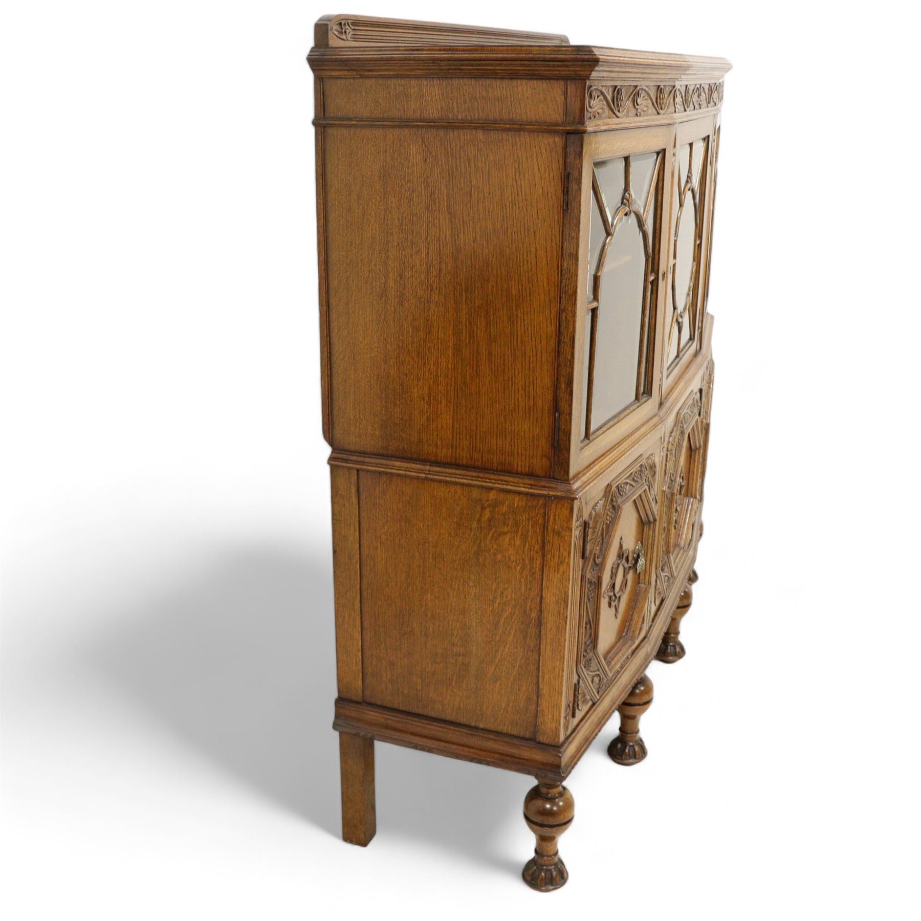 Early 20th century heavily carved oak display cabinet, frieze carved with grape vines over three astragal glazed doors with bevelled glass, over three cupboards applied with octagonal carved panels with further vine carvings, on turned and lobe carved supports