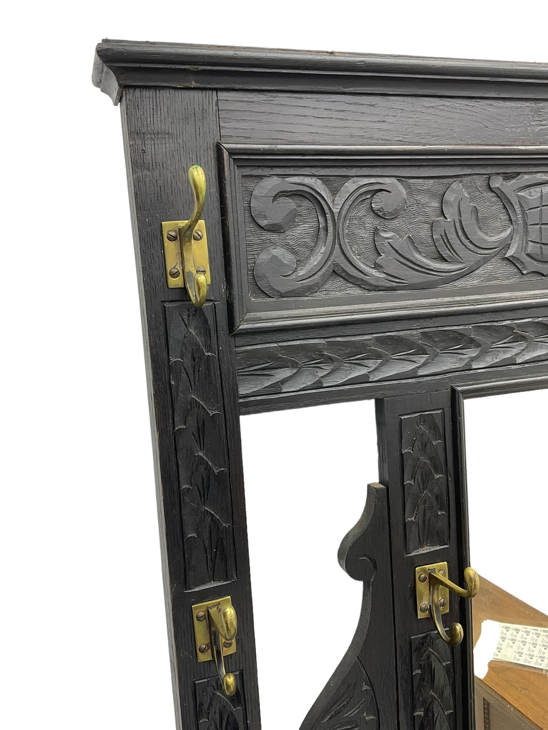 Victorian oak hall stand, the upper section featuring eight brass hooks and central mirror plate flanked by scroll-carved panels, the central section includes a storage box positioned above an umbrella stand, supported by square carved legs with scroll-carved horizontal supports