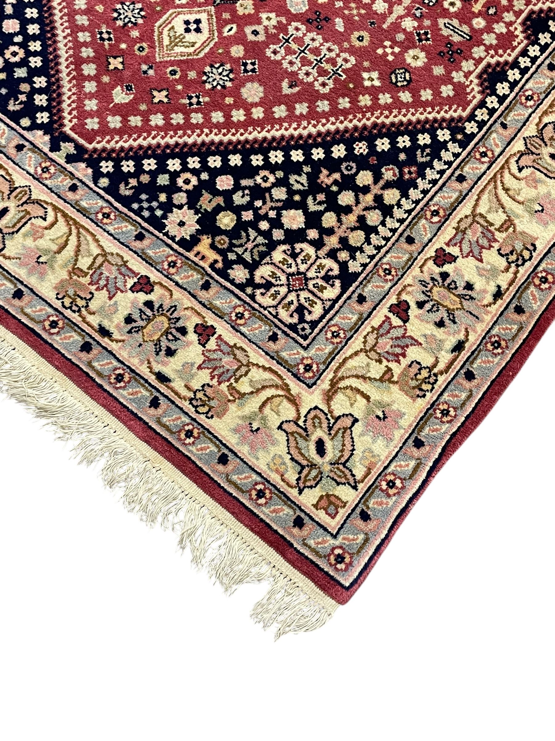 Persian design crimson ground rug, the field decorated with tree of life and floral motifs, enclosed by indigo spandrels with stylised flower head decoration, repeating scrolling border 