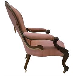 Victorian mahogany framed open armchair, spoon-back and sprung seat upholstered in pink fabric, scrolled arm terminals over serpentine fronted seat decorated with moulded carved floral motifs, cabriole supports with ceramic castors