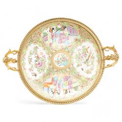 19th century Chinese Canton famille rose circular dish, painted with figures and exotic birds in shaped panels, against a floral and scroll ground, mounted in ormolu style gilt metal twin handled mount with beaded border and three scroll feet, W30cm x H14cm