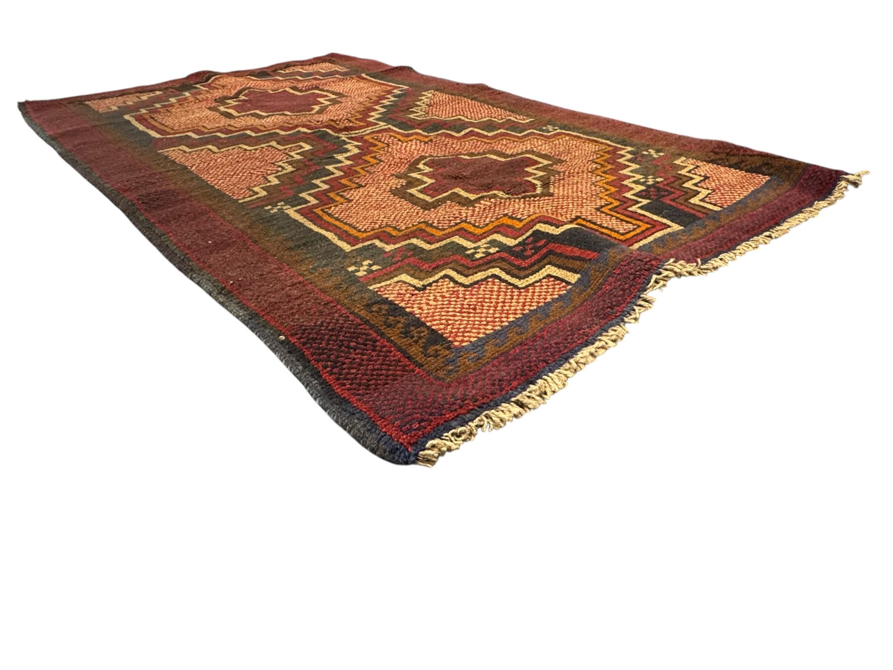 Baluchi crimson ground rug, the central field decorated with two stacked geometric lozenge medallions, surrounded by stepped multicoloured motifs, the wide border with alternating angular motifs in deep red, within a narrow brown guard stripe