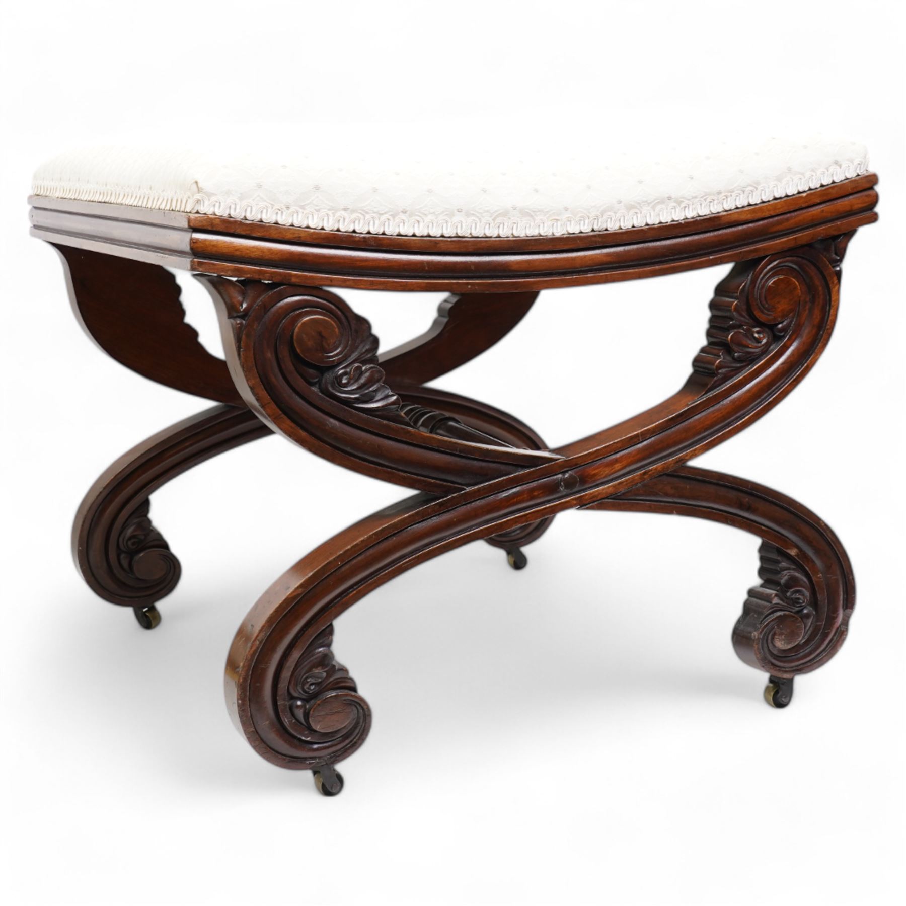 Early 19th century mahogany stool, the dished seat upholstered in cream fabric with repeating pattern, moulded seat rails, on curved X-frame supports carved with scrolls and foliage, united by turned stretcher, on brass castors