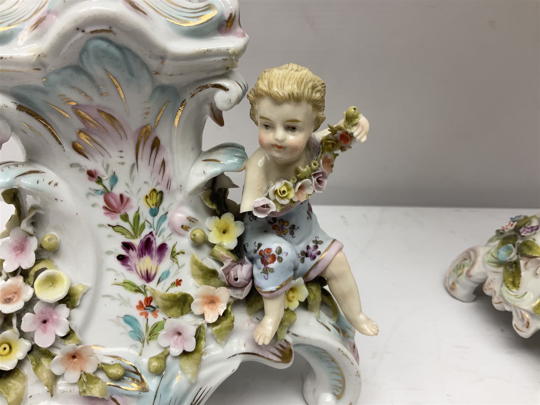 Early 20th century porcelain garniture, the central shaped basket upon a column supported by a female figure, and base with scroll feet, flanked by a pair of four branch candelabra, the scrolling branches and central stem leading to urn shaped sockets over foliate drip pans, upon shaped columns and bases with applied putto, the whole with encrusted flowers throughout, basket H33.5cm, candelabra H52cm