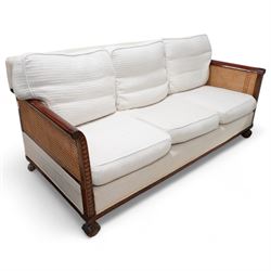 Early 20th century mahogany framed bergère three seat sofa, upholstered back and seat flanked by rolled double-caned arms with carved scale decoration, floral and ribbon-twist carved lower edge, over ball-and-claw feet