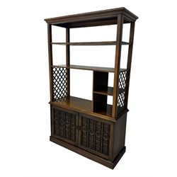 Mid 20th century oak bookcase cupboard, projecting cornice over three open shelves with subdivided sections and latticework uprights, two doors to the base with floral and scrollwork carvings and central brass handles, resting on a moulded plinth base